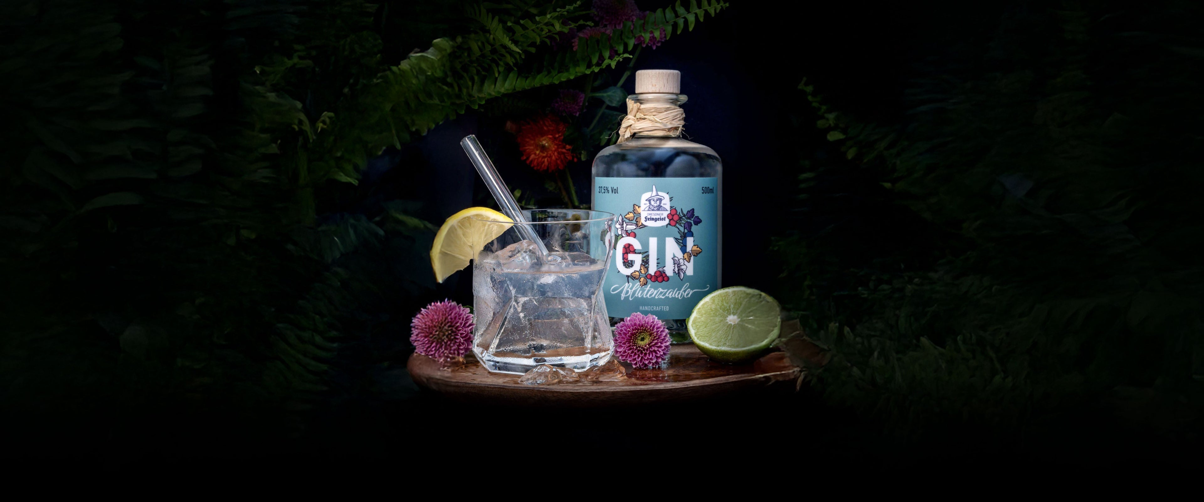Stimmungsvolle Szene mit der Gin-Sorte „Blütenzauber“ von Dresdner Feingeist: Eine blaue Flasche steht neben einem Glas mit Eiswürfeln, Zitronenscheibe und Metallstrohhalm. Umgeben von Farnblättern, lila Distelblüten und einer aufgeschnittenen Limette entsteht eine natürliche, elegante Atmosphäre.