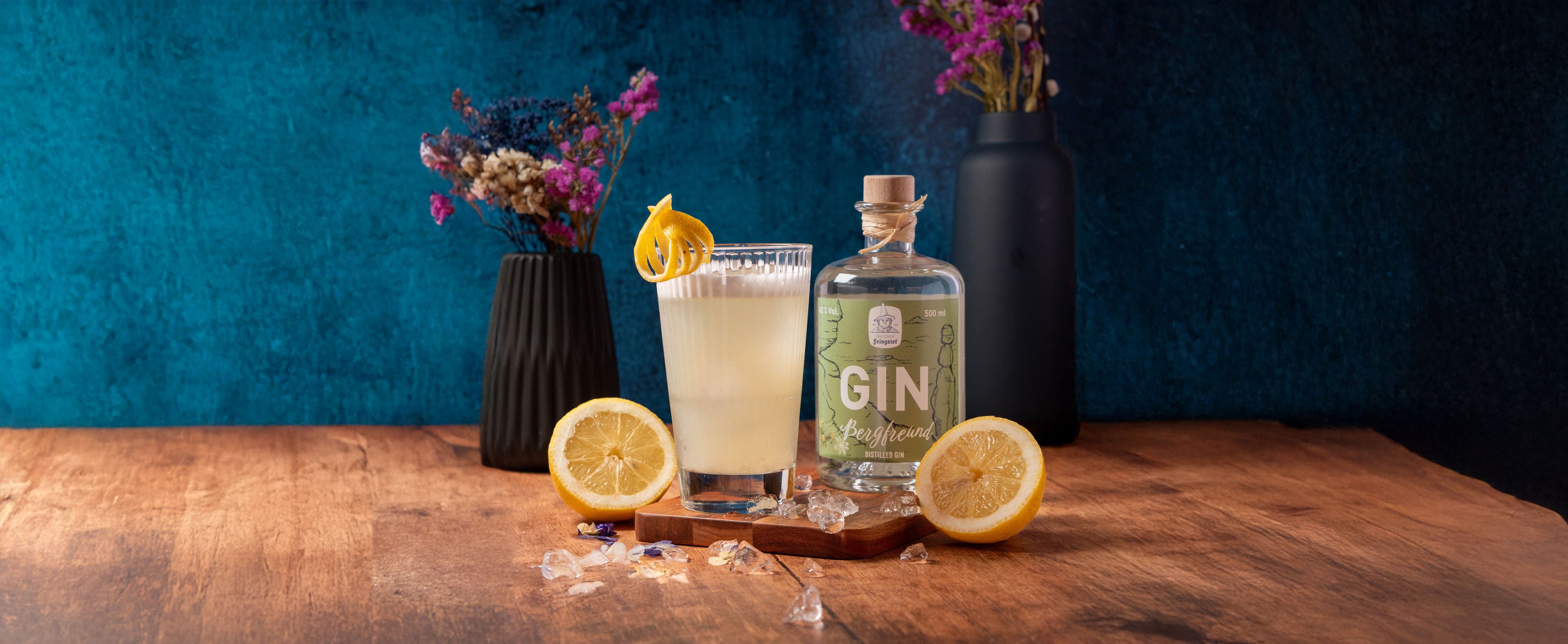 Ein cremiger, zitronengelber Gin-Cocktail mit Schaumkrone und dekorativer Zitronenzeste steht auf einem Holzbrett neben einer Flasche Dresdner Feingeist Gin „Bergfreund“. Eiswürfel und halbierte Zitronen liegen auf dem Holztisch, im Hintergrund kontrastieren dunkle Vasen mit Trockenblumen das szenische Arrangement vor einer tiefblauen Wand.
