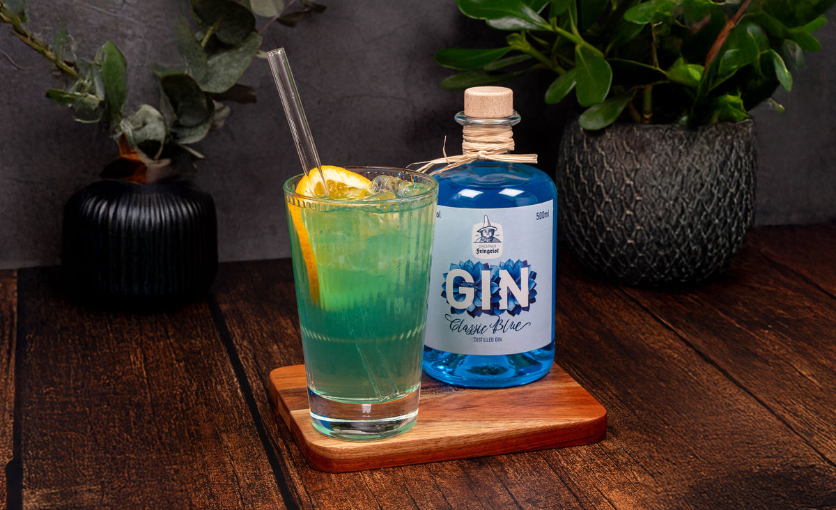 Grünlich schimmernder Gin-Longdrink im Highballglas mit Zitronenzeste und Glasstrohhalm vor einer Flasche Dresdner Feingeist GIN Classic Blue, arrangiert auf Holzbrett mit Pflanzen im Hintergrund auf dunklem Holztisch in eleganter Studioatmosphäre.