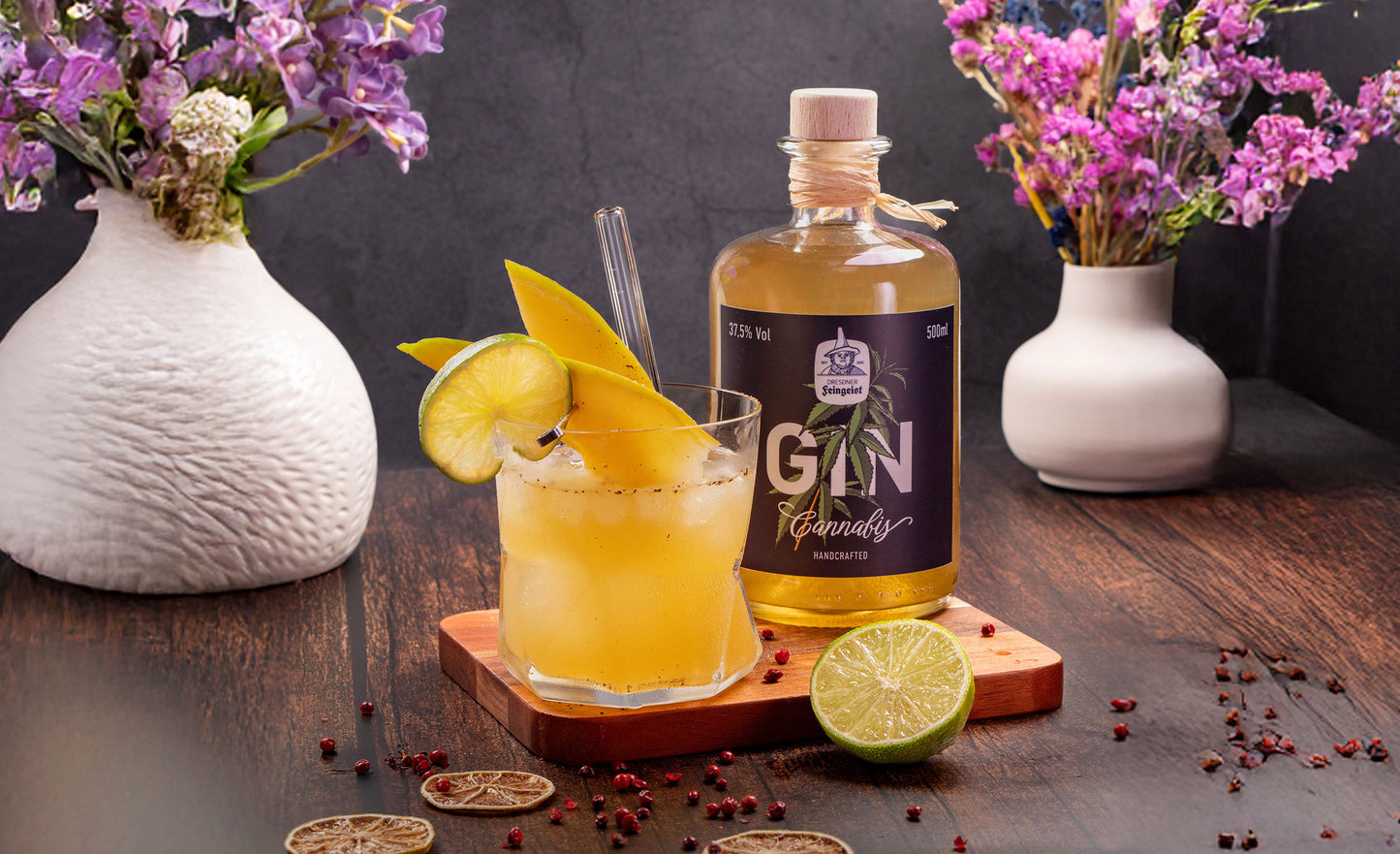 Ein leuchtend gelber Gin-Cocktail, garniert mit Mangoscheiben und einer Limettenspalte, steht auf einem Holzuntersetzer. Eine Flasche Dresdner Feingeist Gin Cannabis ist daneben platziert. Auf dem dunklen Holztisch liegen getrocknete Limettenscheiben und rosa Pfefferbeeren verstreut. Im Hintergrund sind zwei weiße Vasen mit lila und rosa Blumen zu sehen.
