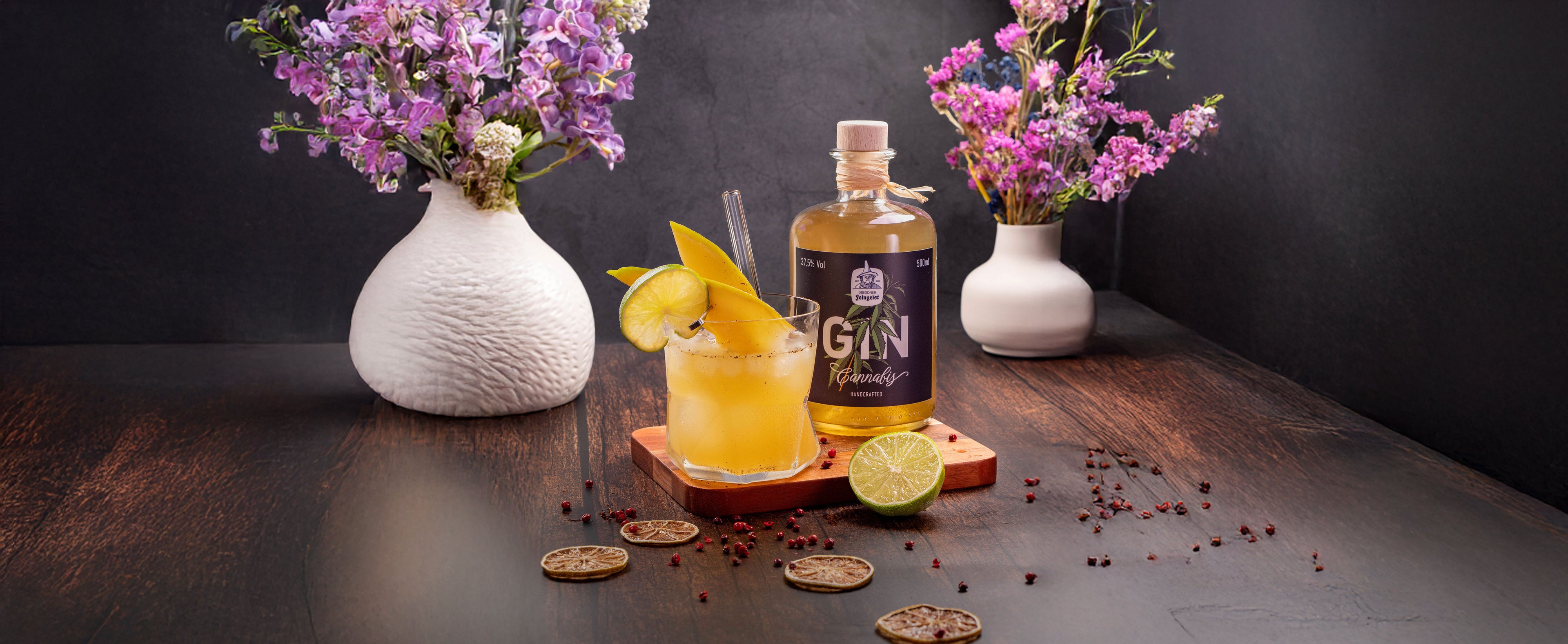 Ein leuchtend gelber Gin-Cocktail, garniert mit Mangoscheiben und einer Limettenspalte, steht auf einem Holzuntersetzer. Eine Flasche Dresdner Feingeist Gin Cannabis ist daneben platziert. Auf dem dunklen Holztisch liegen getrocknete Limettenscheiben und rosa Pfefferbeeren verstreut. Im Hintergrund sind zwei weiße Vasen mit lila und rosa Blumen zu sehen.