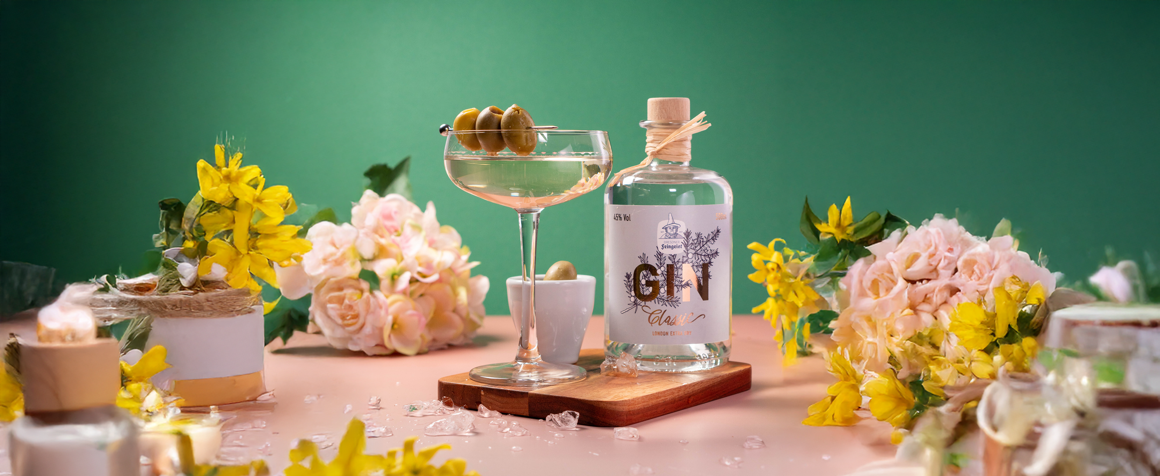 Martini-Cocktail mit grünen Oliven im Coupéglas neben einer 500 ml Flasche Dresdner Feingeist Classic Gin, umgeben von Blumen auf rosa Untergrund mit grünem Hintergrund.