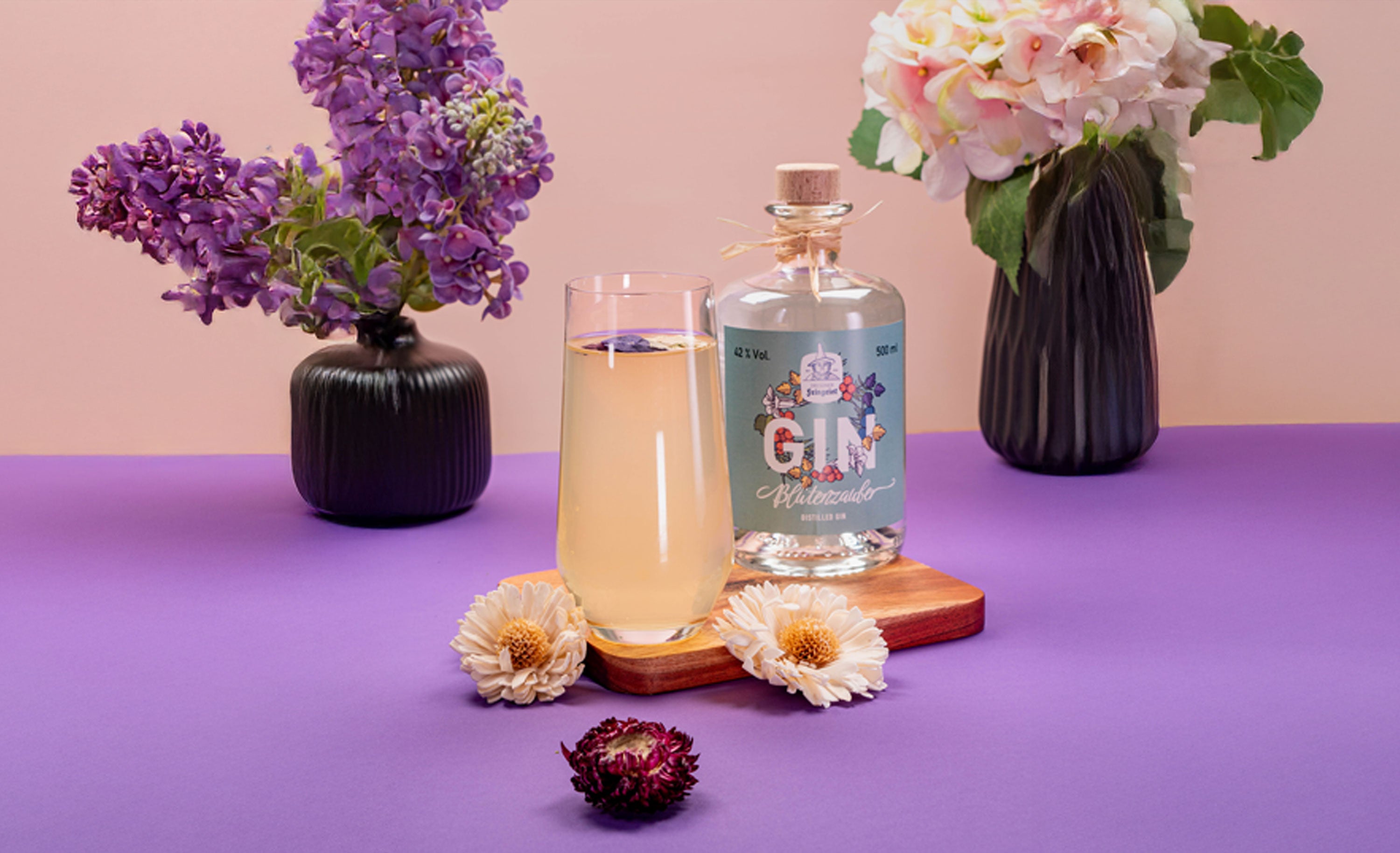 Ein eleganter Cocktail in einer Champagnerflöte, serviert mit der Dresdner Feingeist Blütenzauber Ginflasche auf einem Holzbrett. Der Drink steht auf einem violetten Untergrund, umgeben von Trockenblumen und dekorativen Vasen mit Flieder- und Hortensienblüten – ein frischer, floraler Look im Frühlingsstil.