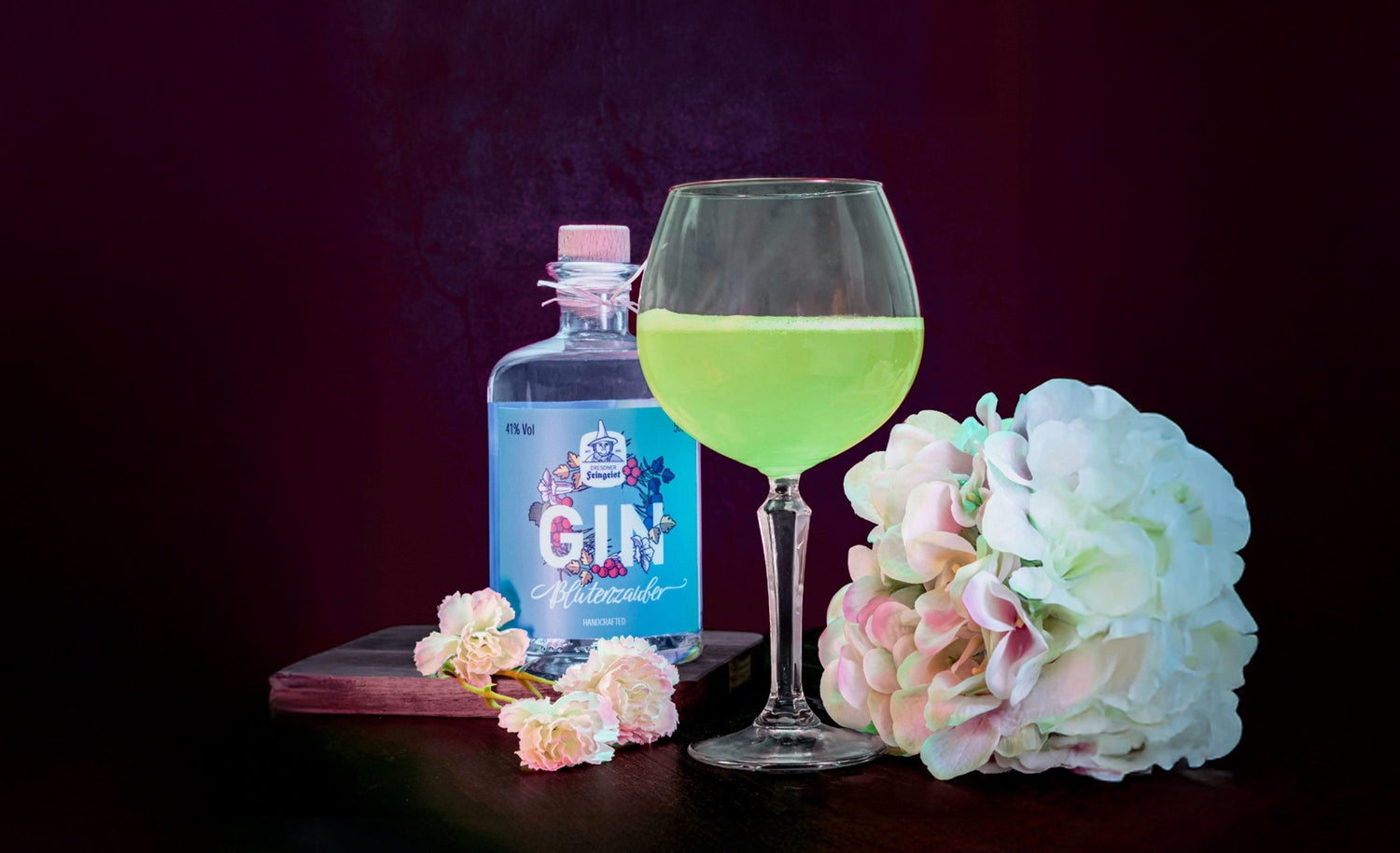 Ein leuchtend grüner Cocktail in einem bauchigen Weinglas steht auf einem dunklen Tisch neben einer Flasche Dresdner Feingeist Blütenzauber Gin. Weiße und zartrosa Blüten umrahmen das Arrangement, das vor einem tief dunkelroten Hintergrund stilvoll in Szene gesetzt ist.