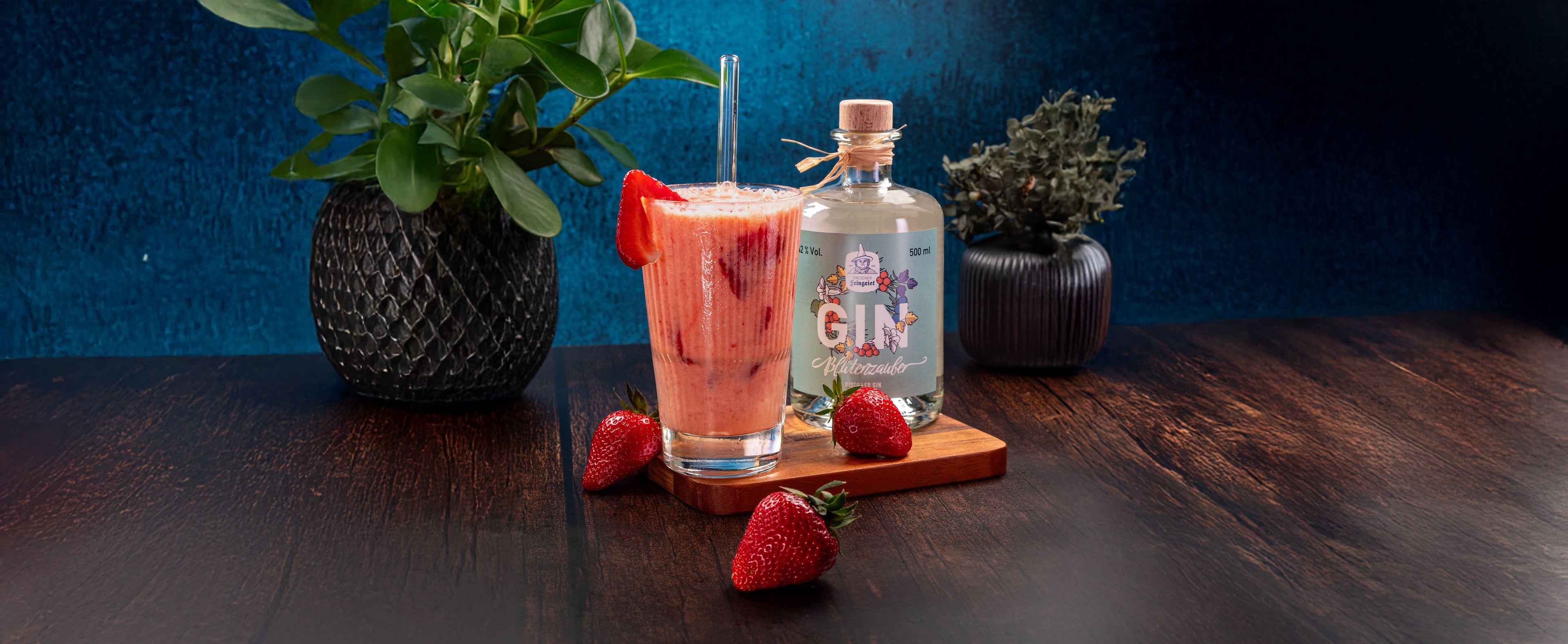 Ein sommerlicher Erdbeer-Cocktail mit Dresdner Feingeist Blütenzauber Gin in einem hohen Glas mit Strohhalm und frischer Erdbeergarnitur. Umgeben von mehreren frischen Erdbeeren und einer stilvollen Ginflasche auf einem Holzbrett, mit Pflanzenvasen im Hintergrund auf dunklem Holztisch und kräftig blauem Hintergrund.