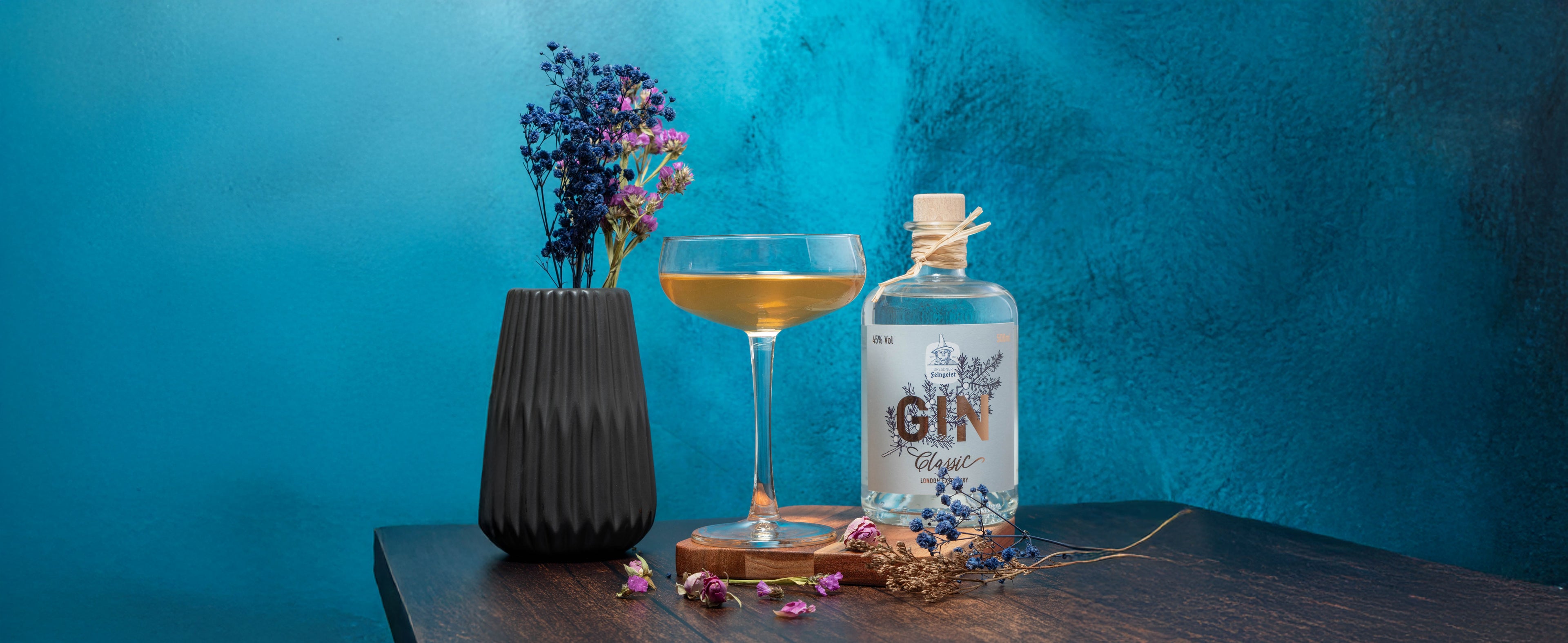 Ein eleganter, bernsteinfarbener Cocktail in einer Coupette steht auf einem Holzbrett neben einer Flasche Dresdner Feingeist Classic Gin. Umgeben ist das Arrangement von blauen Trockenblumen in einer schwarzen Vase und verstreuten Blüten auf einem dunklen Holztisch. Der Hintergrund zeigt eine intensive, strukturierte blaue Wand.