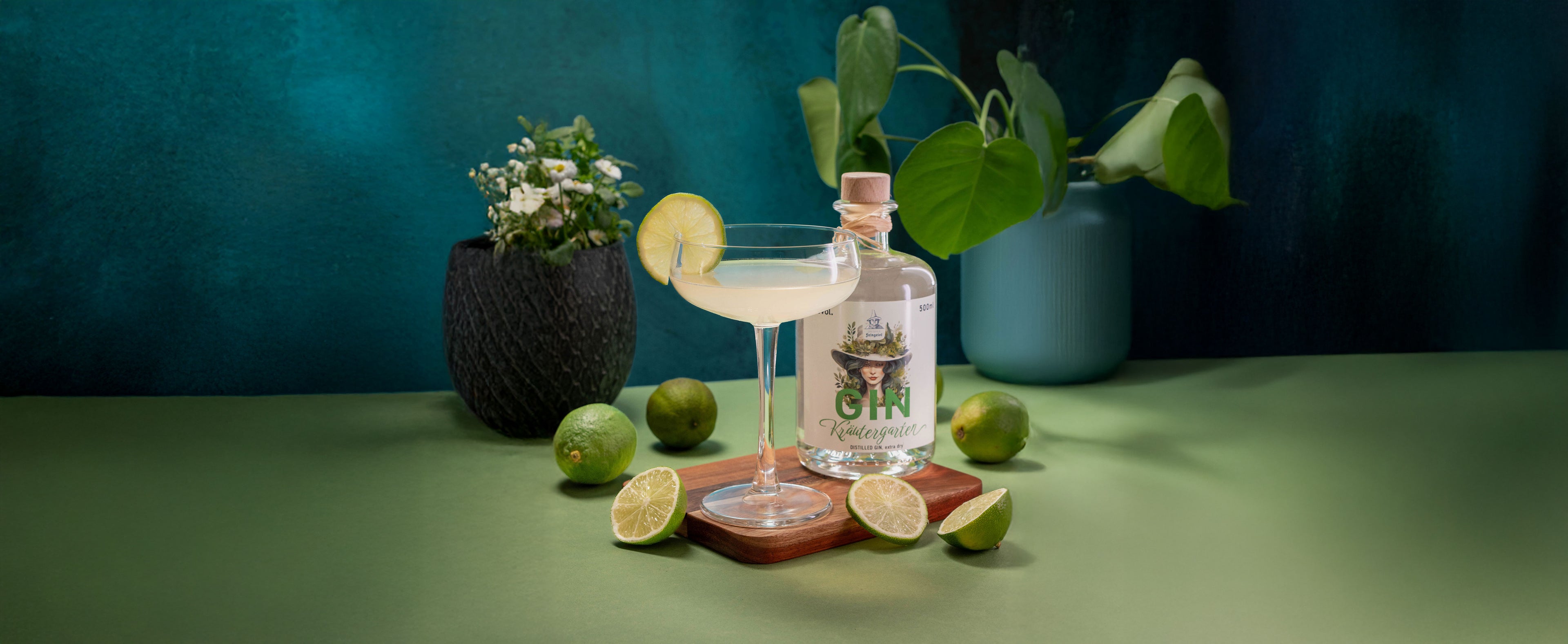 Ein blassgrüner Cocktail in einem Martini-Glas, garniert mit einer Limettenscheibe, steht auf einem Holzuntersetzer. Daneben ist eine Flasche Dresdner Feingeist Gin "Kindergarten" platziert, umgeben von ganzen und halbierten Limetten. Im Hintergrund sind zwei Pflanzen in dunklen und hellblauen Töpfen vor einer dunkelgrünen Wand zu sehen.