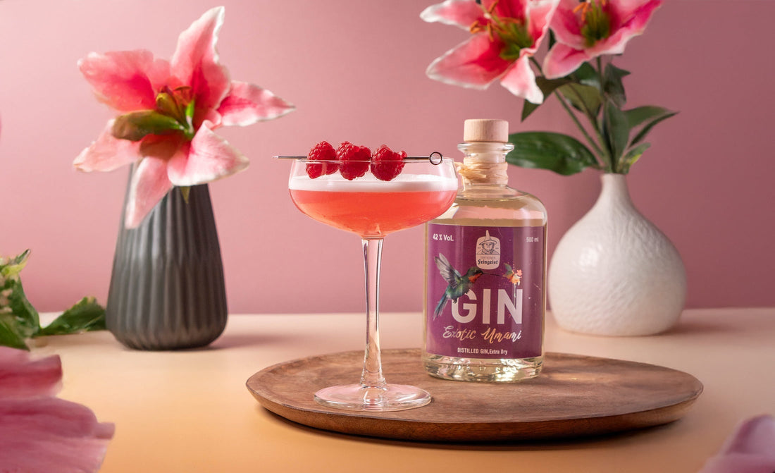 Clover Club Cocktail mit rosa Schaum und frischen Himbeeren auf einem Cocktailspieß, serviert in einer Cocktailschale auf Holztablett – daneben eine Flasche Dresdner Feingeist Exotic Umami Gin, dekoriert mit rosa Lilien im Hintergrund.