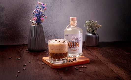 Gin Espresso Martini in einem Tumbler Glas mit Espresso-Schaum, garniert mit Kaffeebohnen, daneben eine Flasche Dresdner Feingeist Classic Gin auf einem Holzbrett.