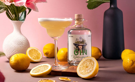 Ein eleganter Cocktail in einer Coupette mit cremigem Schaum, neben einer Flasche Dresdner Feingeist Gin Kräutergarten. Umgeben von frischen und getrockneten Zitronenscheiben, gelben Zitronen und zarten Blumenarrangements auf rosafarbenem Hintergrund.