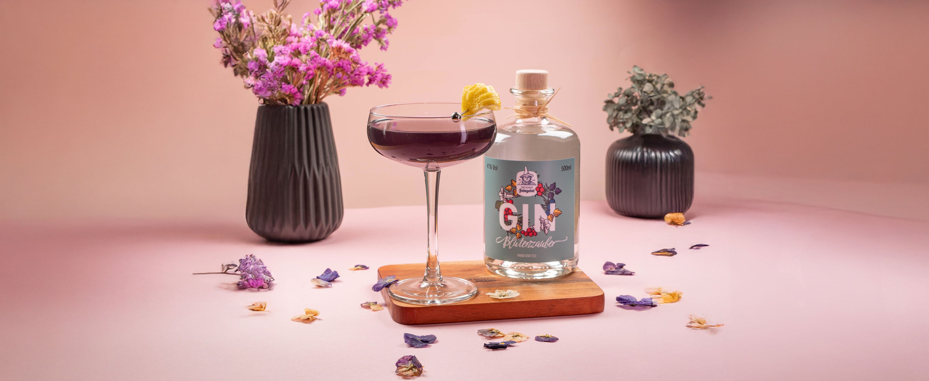 Lila-blauer Cocktail in einer Coupette mit getrockneter Zitrone und Beere garniert, serviert neben einer Flasche Dresdner Feingeist Blütenzauber Gin. Umgeben von Trockenblumen und stilvollen Vasen auf einem zartrosafarbenen Hintergrund.