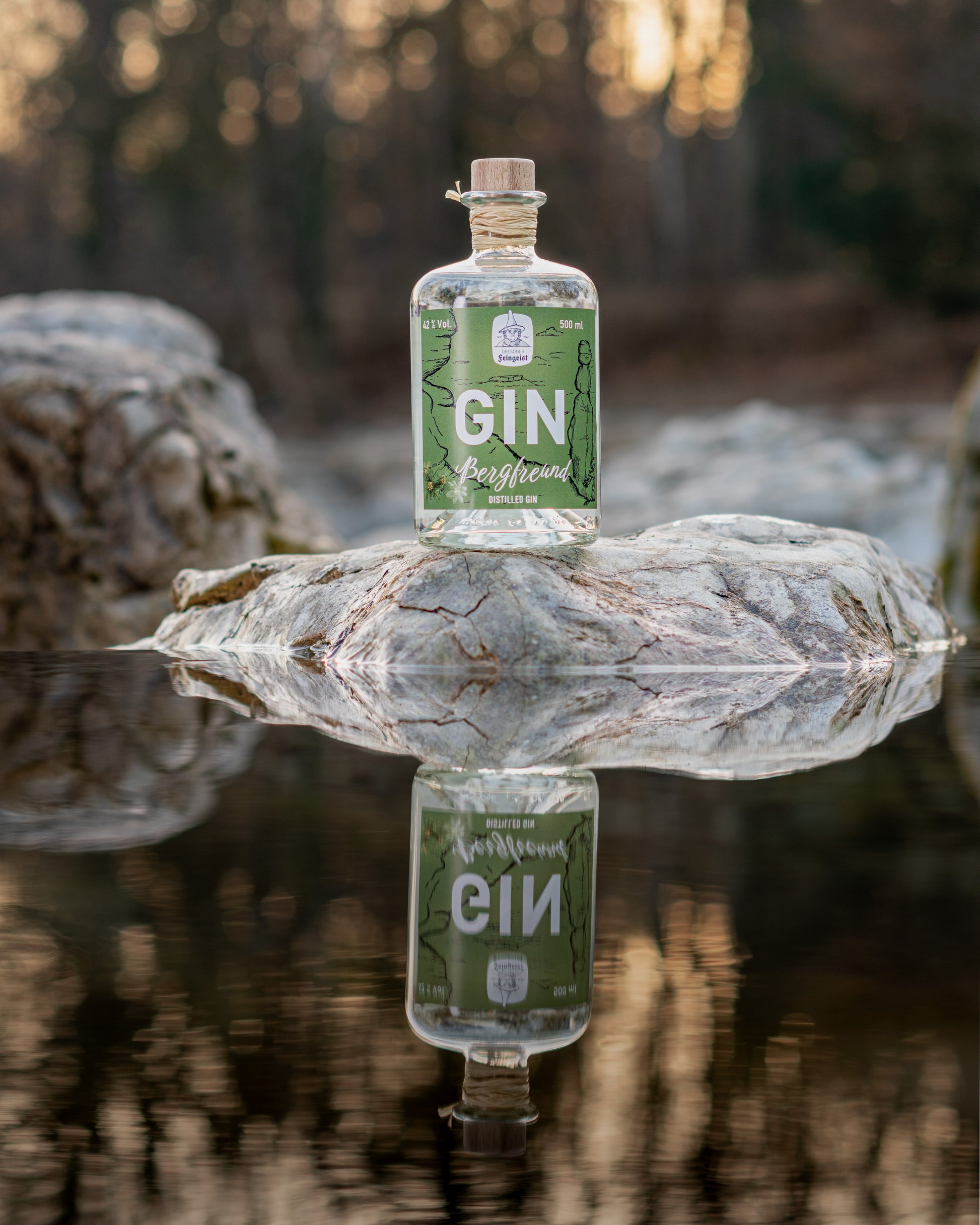 Eine Flasche Dresdner Feingeist Bergfreund steht auf einem Stein über einem kleinen See. Im Wasser spiegelt sich die Flasche beim Sonnenaufgang
