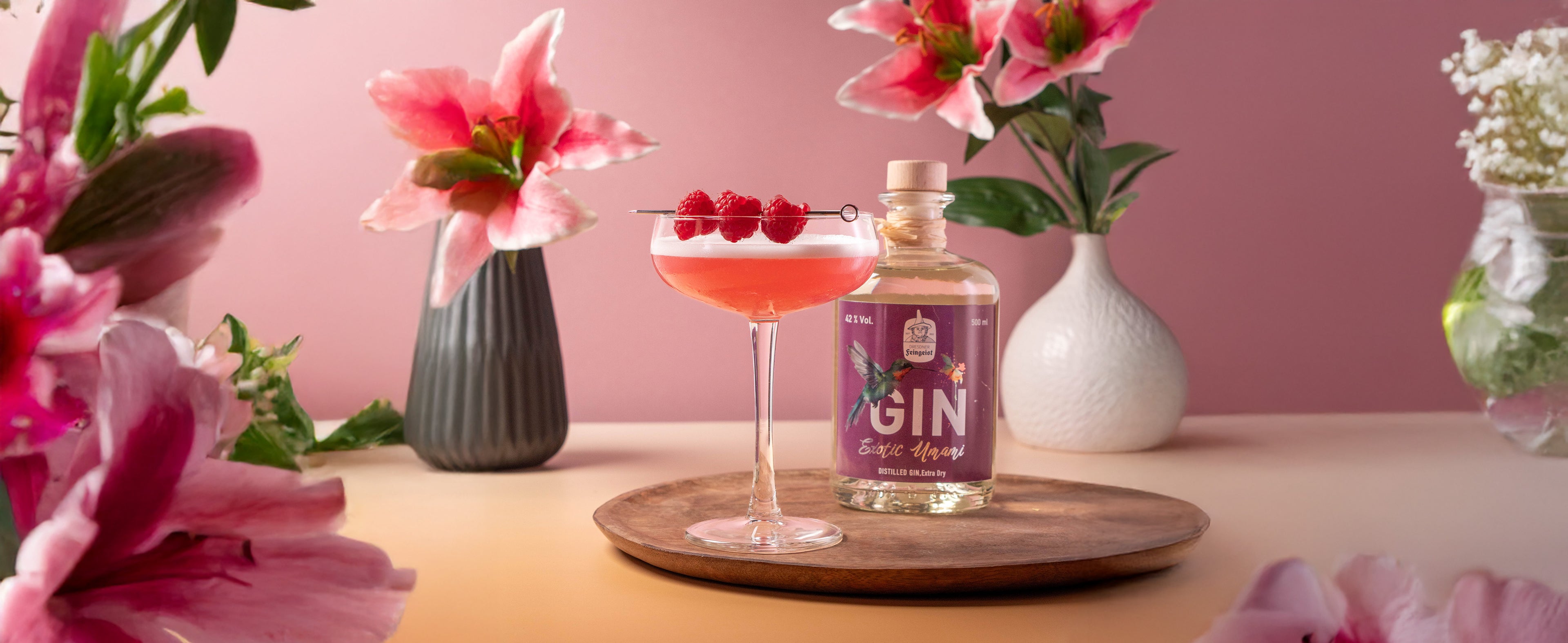 Clover Club Cocktail mit rosa Schaum und frischen Himbeeren auf einem Cocktailspieß, serviert in einer Cocktailschale auf Holztablett – daneben eine Flasche Dresdner Feingeist Exotic Umami Gin, dekoriert mit rosa Lilien im Hintergrund.

