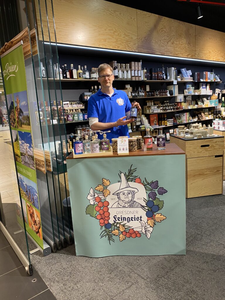 Verkaufsstand von Dresdner Feingeist im Elbepark bei DDV Lokal – ein Mann in blauem Shirt präsentiert eine Ginflasche, der Stand ist mit verschiedenen Gins und Botanicals bestückt und befindet sich in einem gut sortierten Feinkost- und Spirituosenbereich.