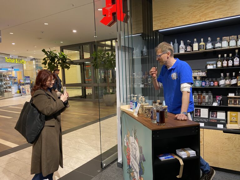 Gin-Verkostung im Elbepark Dresden – ein Mann im blauen Dresdner-Feingeist-Shirt steht hinter dem Probierstand, während eine Frau in Mantel und mit Tasche ein Foto macht. Im Hintergrund ist die Ladenzeile mit Tabak- und Spirituosengeschäft zu sehen.