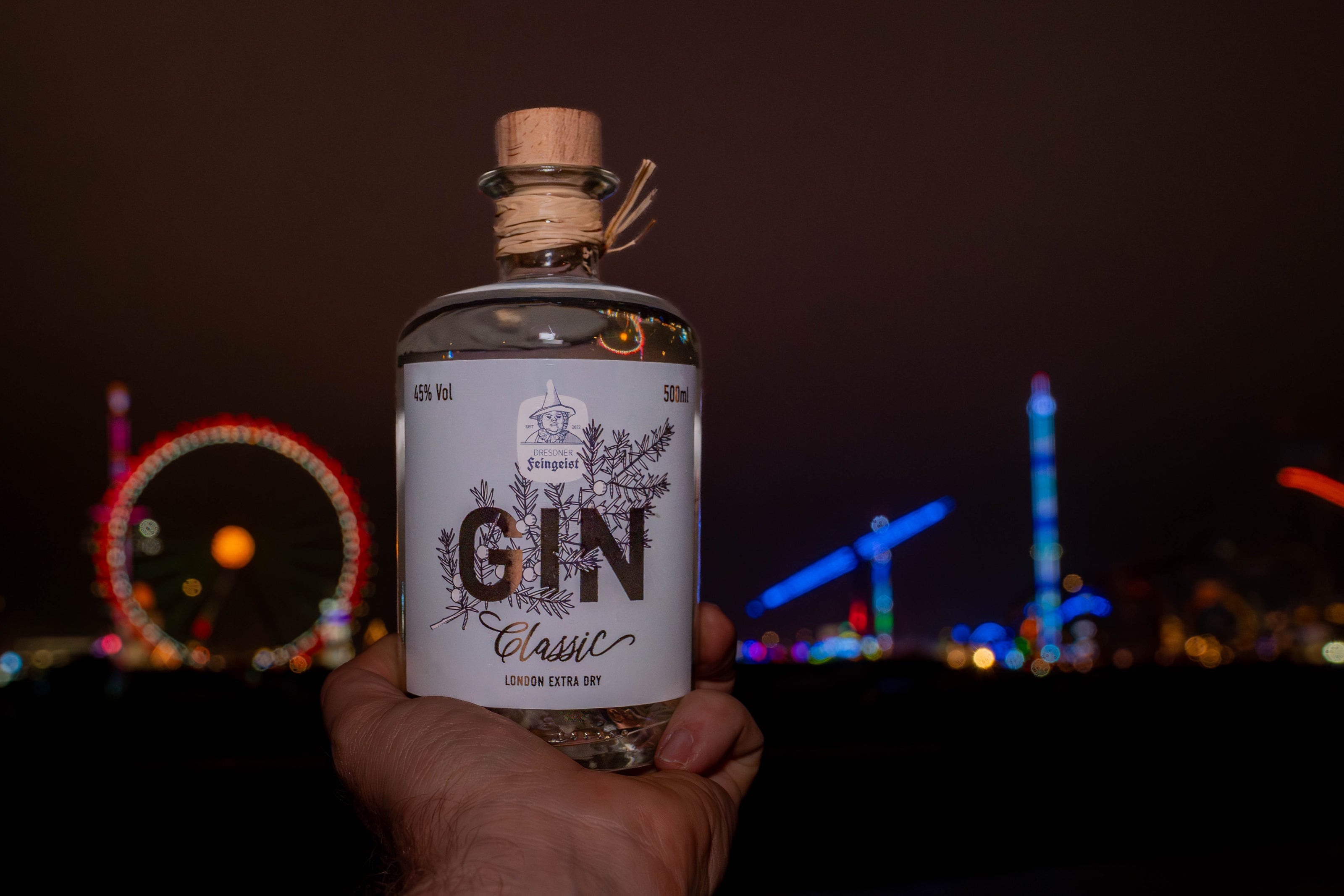Hand hält eine Flasche Dresdner Feingeist GIN Classic – London Extra Dry im Vordergrund, im Hintergrund unscharf beleuchtete Fahrgeschäfte und Lichter der Münchner Wiesn bei Nacht, die eine festliche, urbane Stimmung erzeugen.