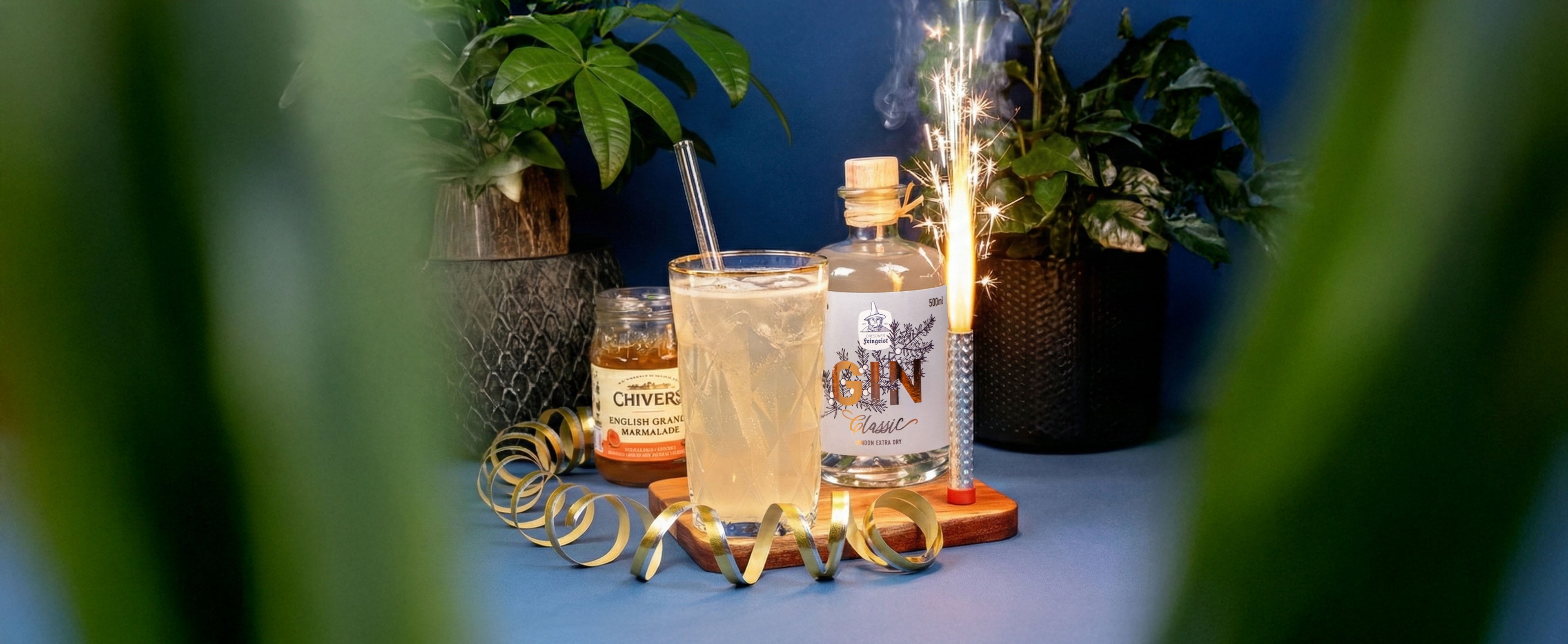 Festlicher Longdrink mit hellem Gin-Cocktail im Highballglas vor einer Flasche Dresdner Feingeist GIN Classic, dekoriert mit Wunderkerze, goldenem Luftschlangenband und Pflanzen im Hintergrund auf blauem Untergrund.