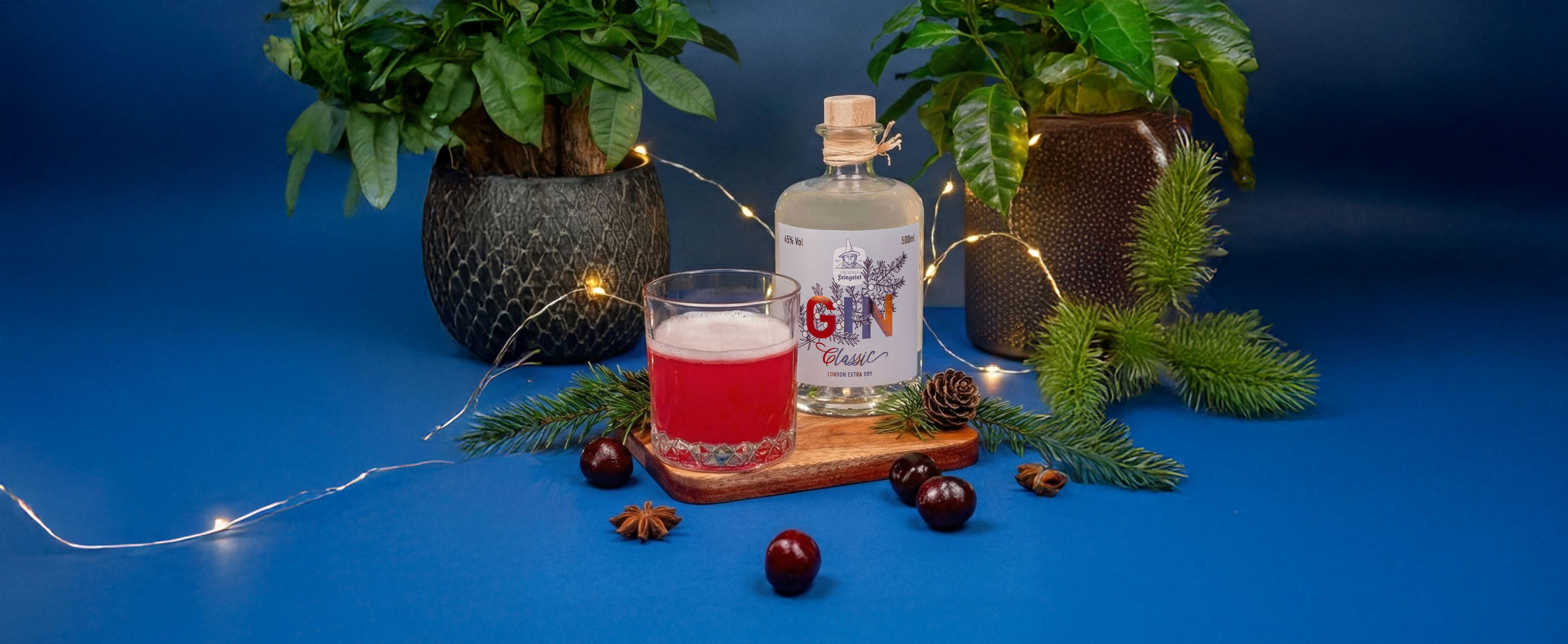 Stillleben mit rotem Winter-Gin-Cocktail im Tumbler neben einer Flasche Dresdner Feingeist GIN Classic, dekoriert mit Tannenzweigen, Gewürzen, Beeren und warmen Lichterketten vor blauem Hintergrund.
