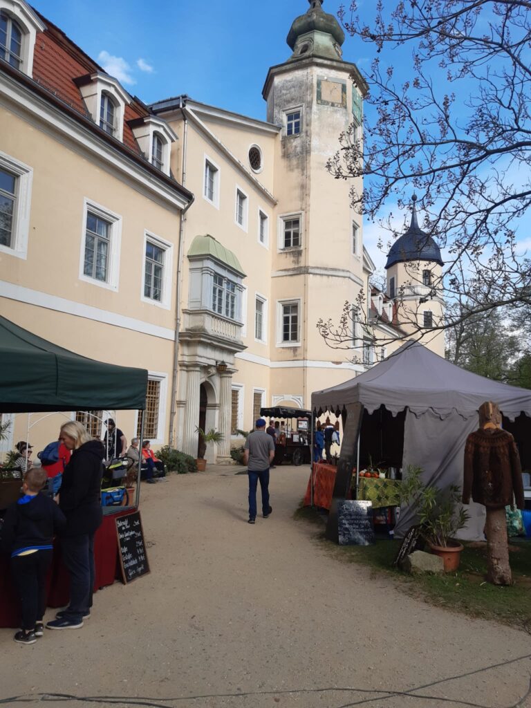 Hoffest auf Schloss Hermsdorf – Marktstände mit Besuchern im Innenhof des Schlosses, darunter ein Verkaufszelt für regionale Produkte. Im Hintergrund das helle Schlossgebäude mit Türmen und blauem Himmel.