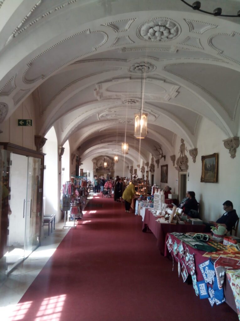 ALT-Text: Verkaufsstände im barocken Innenflur von Schloss Hermsdorf – unter reich verziertem Gewölbe präsentieren verschiedene Aussteller Kunsthandwerk, Kleidung und regionale Produkte entlang eines roten Teppichs.