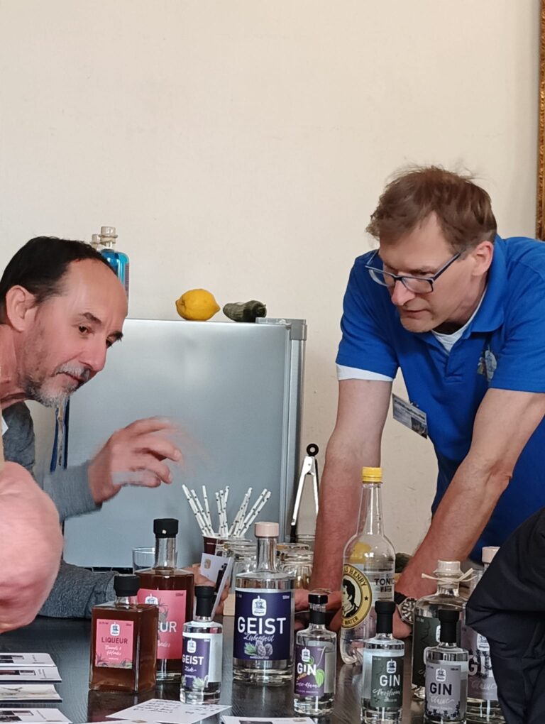 Zwei Männer im Gespräch an einem Verkostungstisch mit verschiedenen Gin- und Likörflaschen von Dresdner Feingeist; im Hintergrund ein Kühlschrank mit Zitrone und Gurke, Fokus auf Austausch und Beratung.