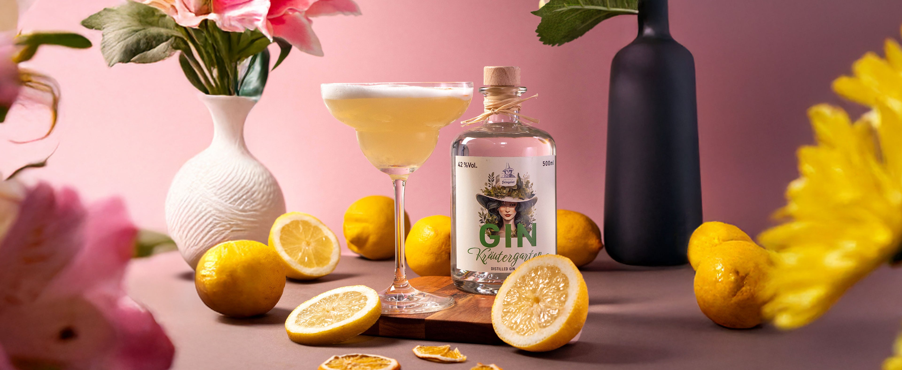 Ein eleganter Cocktail in einer Coupette mit cremigem Schaum, neben einer Flasche Dresdner Feingeist Gin Kräutergarten. Umgeben von frischen und getrockneten Zitronenscheiben, gelben Zitronen und zarten Blumenarrangements auf rosafarbenem Hintergrund.