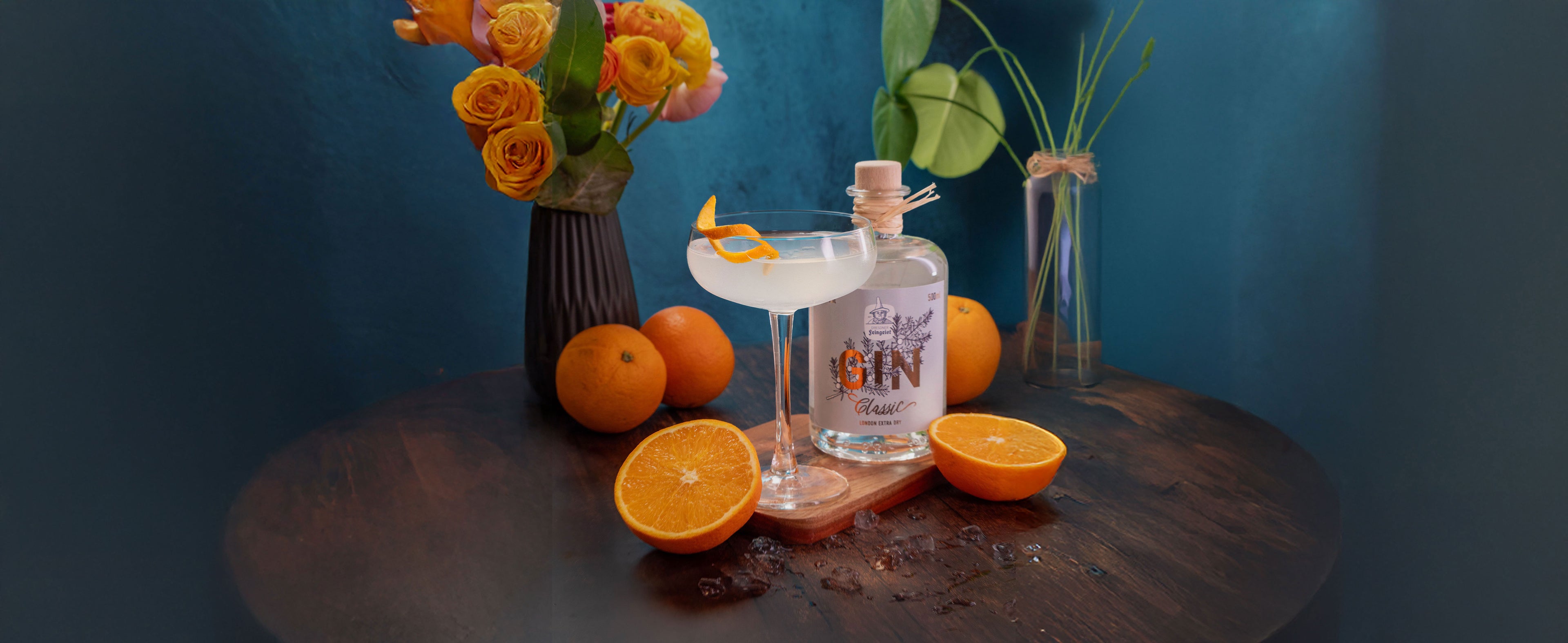 Ein eleganter, heller Cocktail in einem Coupette-Glas, garniert mit einer Orangenzeste, steht auf einem Holzuntersetzer. Daneben befindet sich eine Flasche Dresdner Feingeist Classic Gin, umgeben von ganzen und halbierten Orangen. Im Hintergrund sind ein Strauß orangefarbener Rosen in einer dunklen Vase und eine Pflanze in einer Glasvase auf einem dunklen Holztisch vor einer blauen Wand zu sehen.