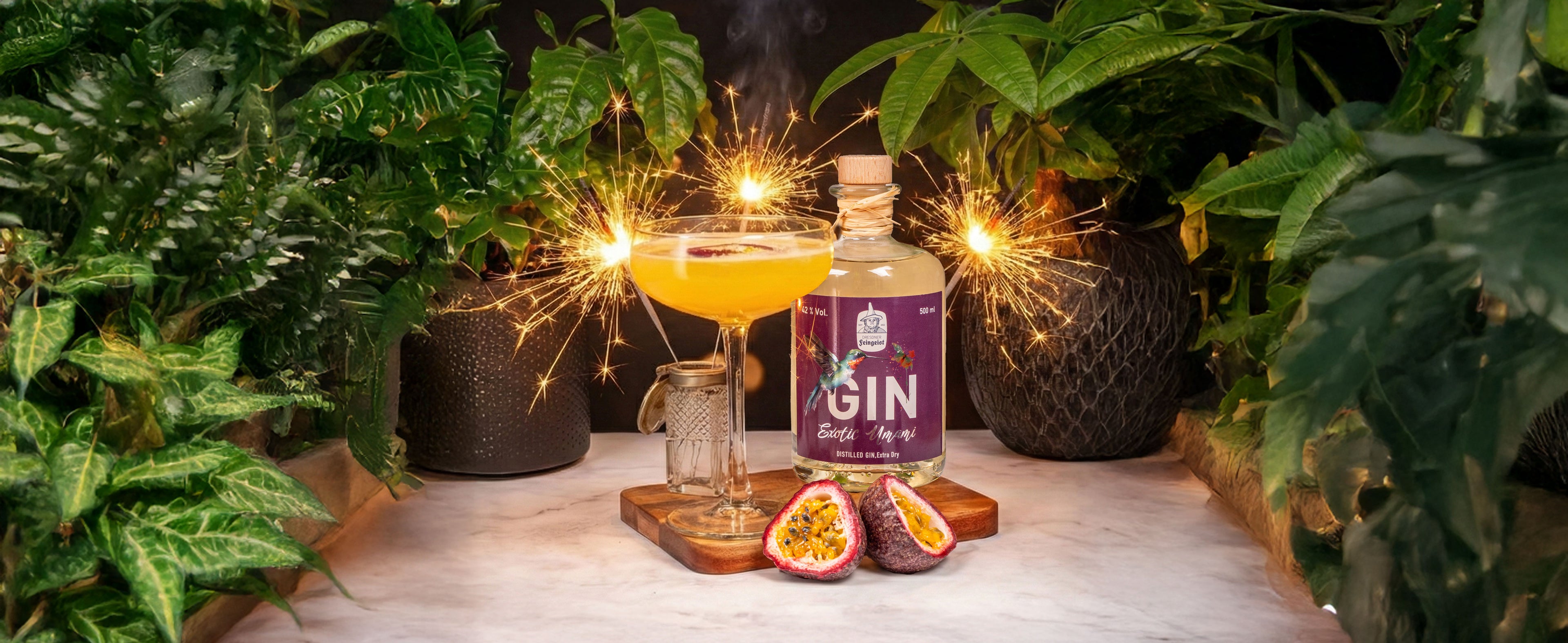 Gelber Gin-Cocktail im Coupeglas mit Wunderkerzen, davor eine halbierte Passionsfrucht, daneben eine Flasche Dresdner Feingeist GIN Exotic Umami auf Holzbrett, inszeniert mit üppigen Pflanzen und warmer, festlicher Atmosphäre.