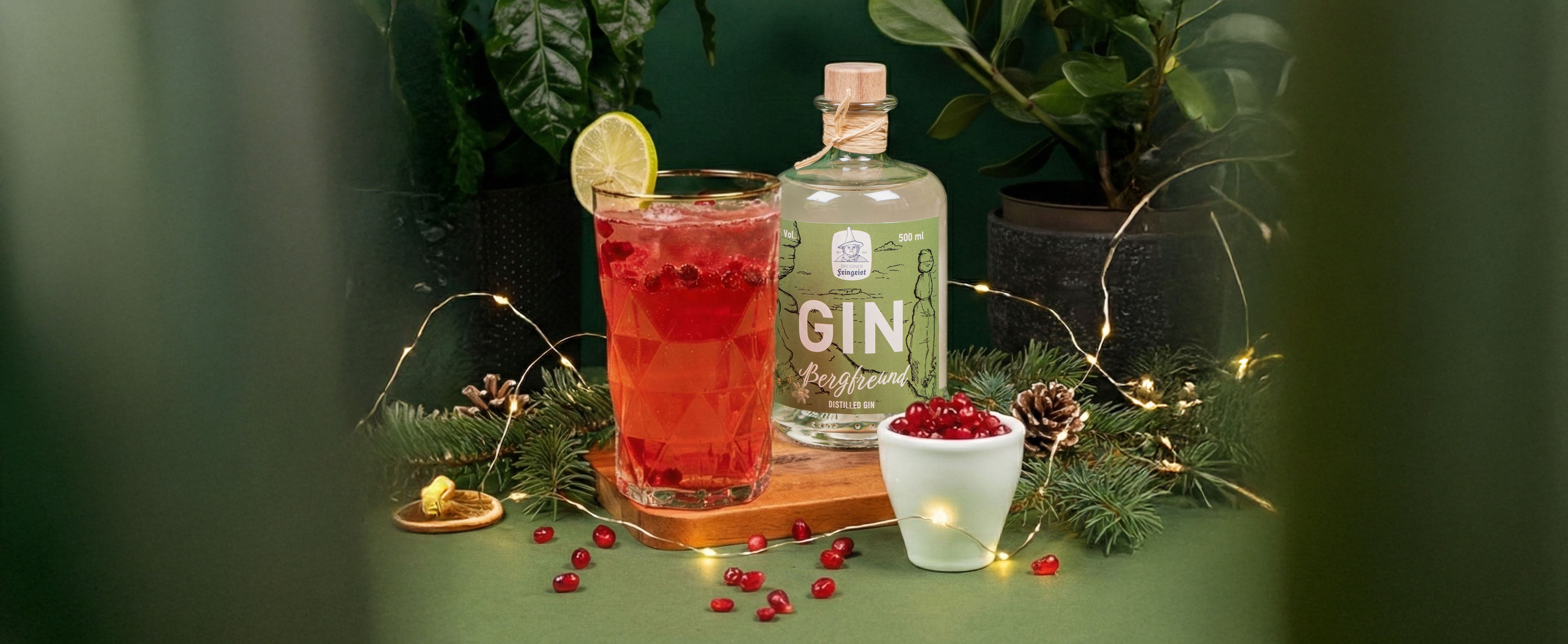 Fruchtiger roter Gin-Longdrink im Tumbler mit Limettenscheibe vor einer Flasche Dresdner Feingeist GIN Bergfreund, arrangiert mit Preiselbeeren, Tannenzweigen, Zapfen und feiner Lichterkette in winterlich-natürlicher Atmosphäre.