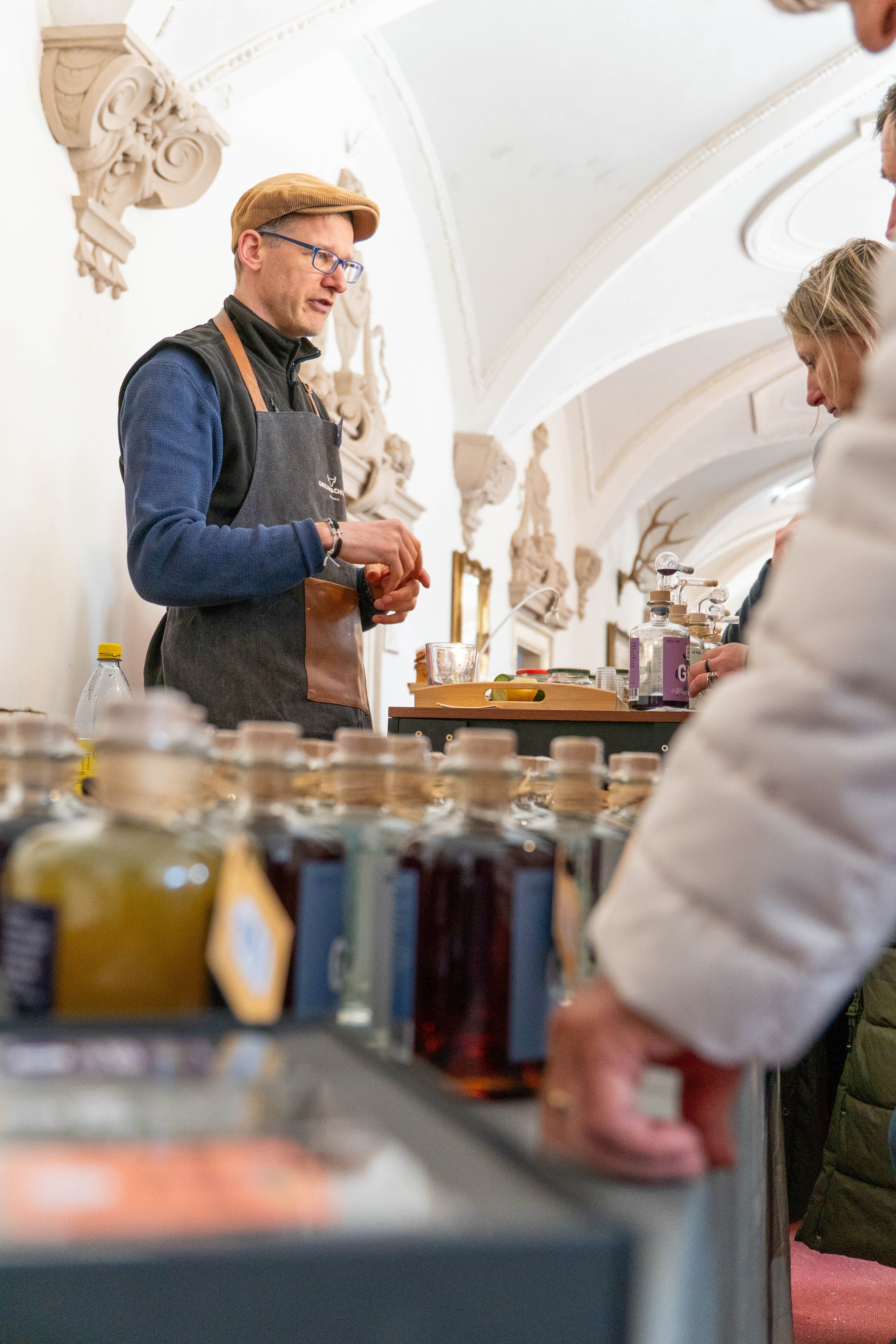 Ein Mann mit Schiebermütze und Schürze steht hinter einem Verkostungsstand von Dresdner Feingeist. Vor ihm sind sechs geöffnete Ginflaschen mit Glas-Ausgießern aufgereiht – darunter die Sorten Classic, Blütenzauber, Bergfreund, Blaues Wunder und Exotic Umami. Im Hintergrund hängt ein gerahmtes Gemälde.