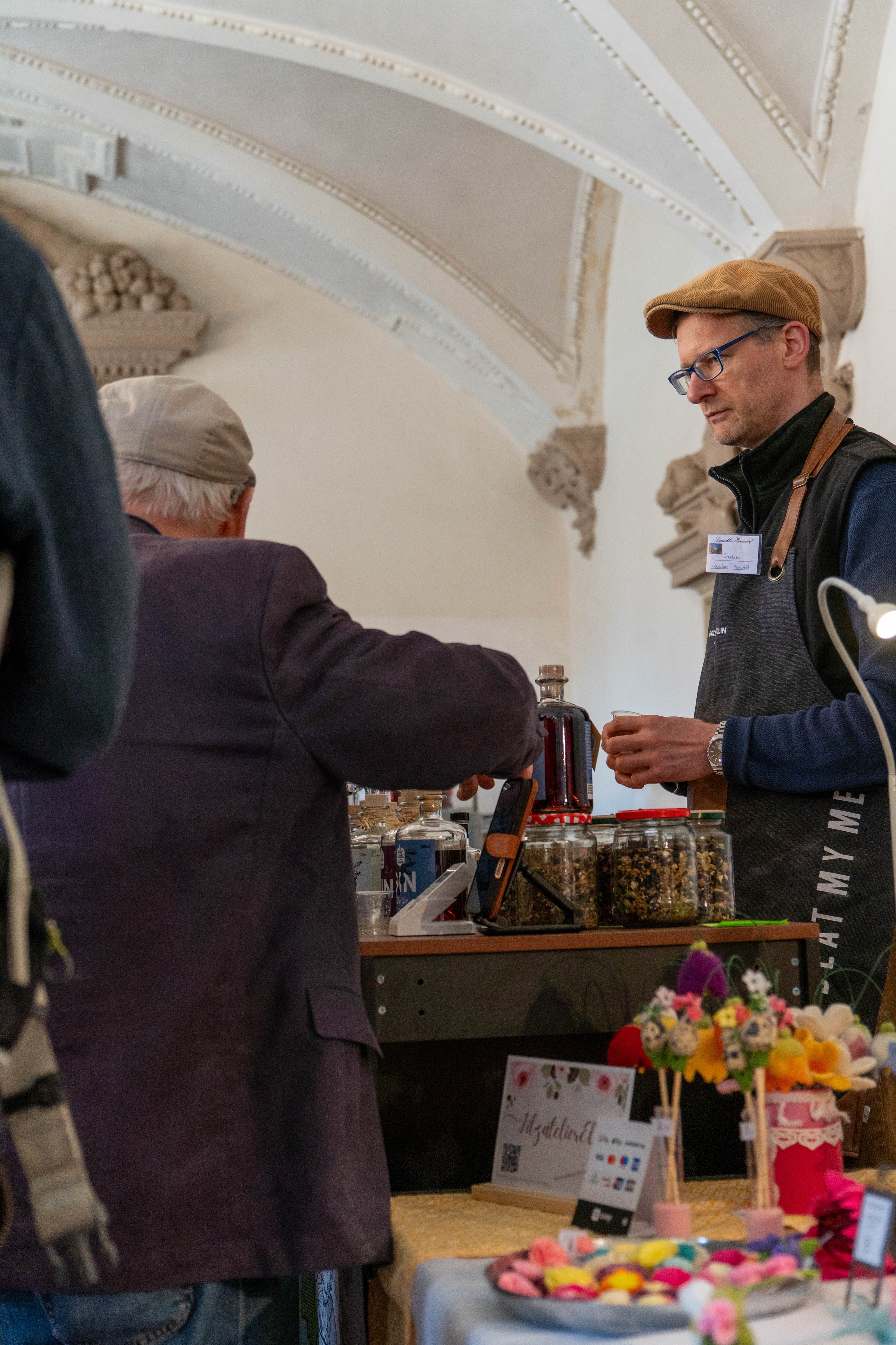Ein Verkäufer mit Schiebermütze und Schürze schenkt einem älteren Mann am Stand von Dresdner Feingeist Gin ein Probierglas ein. Der Stand ist mit Ginflaschen, Kräutergläsern und einem Smartphone bestückt. Im Vordergrund steht ein weiterer Marktstand mit bunten Filzblumen. Die Szene spielt in einem prunkvollen Saal mit hohen Gewölbedecken.
