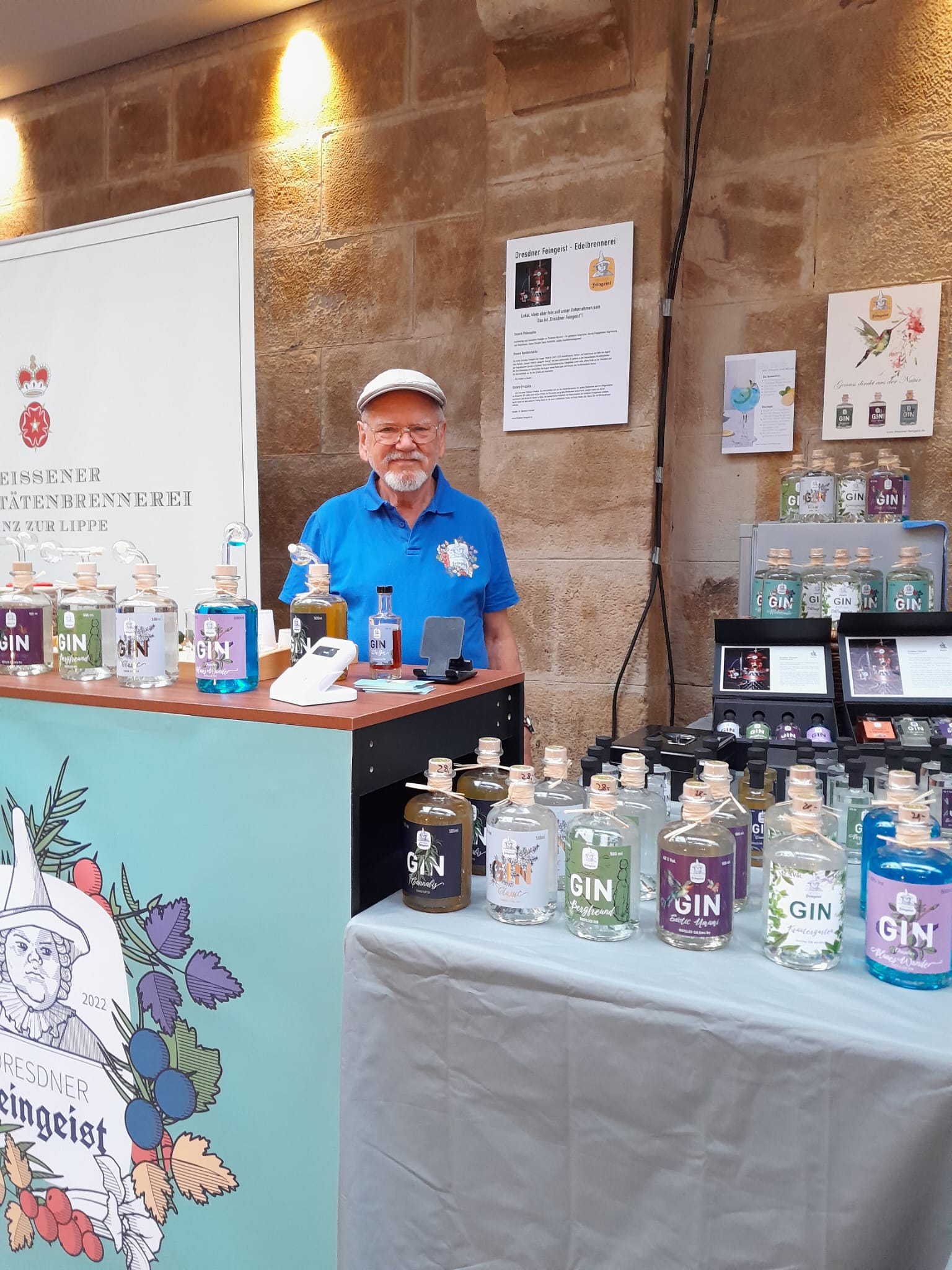 Ein älterer Mann mit weißer Mütze und blauem Polohemd steht an einem Marktstand von „Dresdner Feingeist“. Vor ihm sind viele bunte Ginflaschen ausgestellt. Im Hintergrund ist eine Sandsteinwand mit Postern und Schildern zu sehen. Auf dem Tresen ist das Dresdner-Feingeist-Logo mit historischer Figur und Beeren abgebildet.