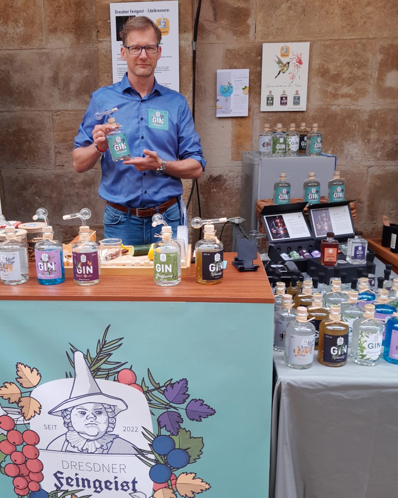 Ein Mann in blauem Hemd steht hinter einem liebevoll gestalteten Marktstand von „Dresdner Feingeist“ und hält eine Flasche „Gin Bergfreund“ in den Händen. Vor ihm stehen zahlreiche Ginsorten mit Glasaufsätzen auf dem Tresen. Rechts daneben befinden sich weitere Flaschen und Probiergläser auf einem Tisch und in einer Präsentationsbox. Der Stand ist mit einem großen Logo der Marke dekoriert, im Hintergrund eine Sandsteinwand mit Plakaten zur Marke.