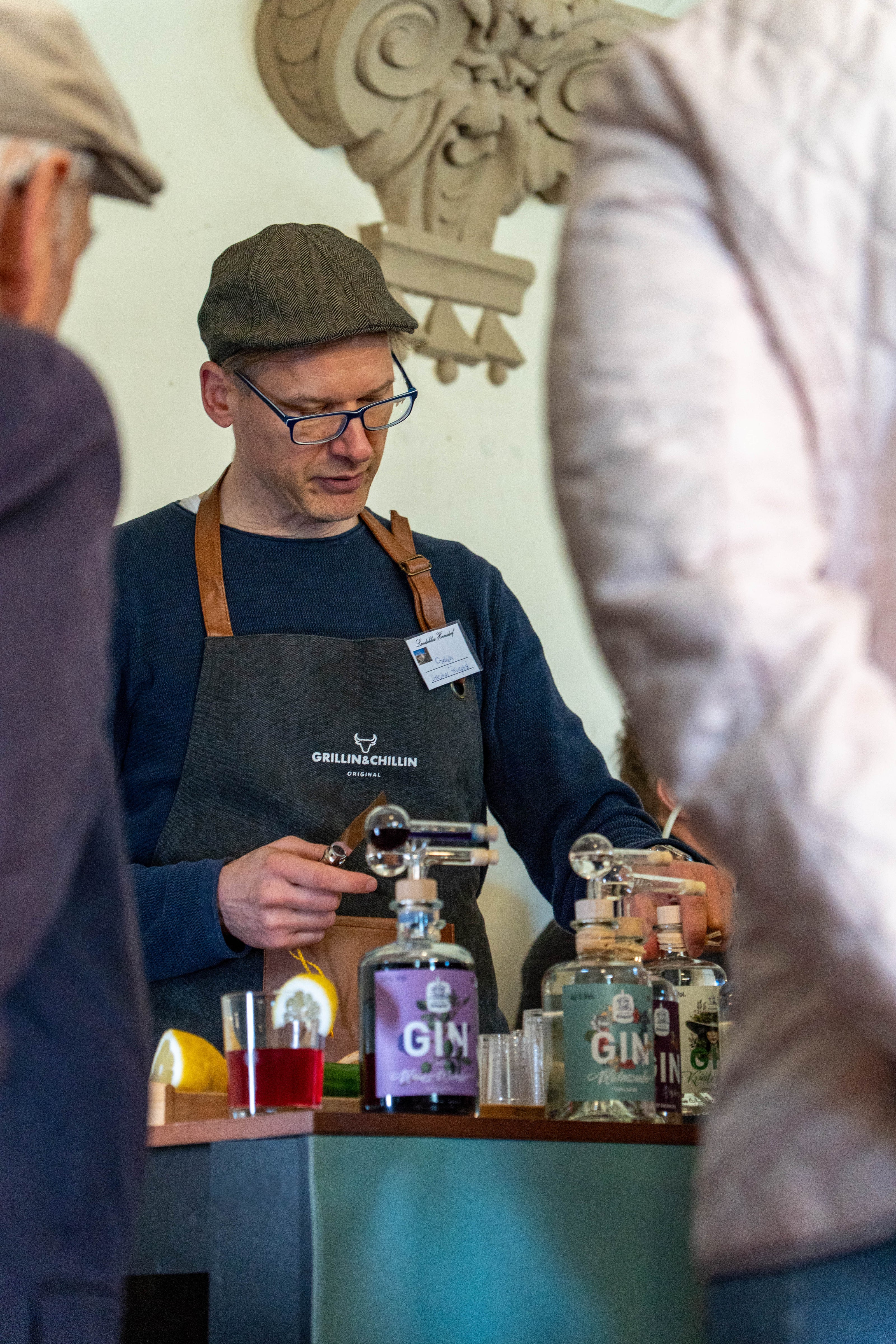 Ein Mann mit Schiebermütze, Brille und dunkelblauer Kleidung steht hinter einem Stand von Dresdner Feingeist. Er trägt eine dunkelgraue „GRILLIN & CHILLIN“-Schürze und bereitet mit Konzentration einen Drink zu. Auf dem Tisch stehen verschiedene Ginflaschen, darunter “Blütenzauber”, “Bergfreund” und “Cannabis Gin”, sowie vorbereitete Cocktailzutaten. Die Szene spielt sich auf einem Hoffest oder Markt ab, weitere Personen sind im Vordergrund angeschnitten sichtbar.