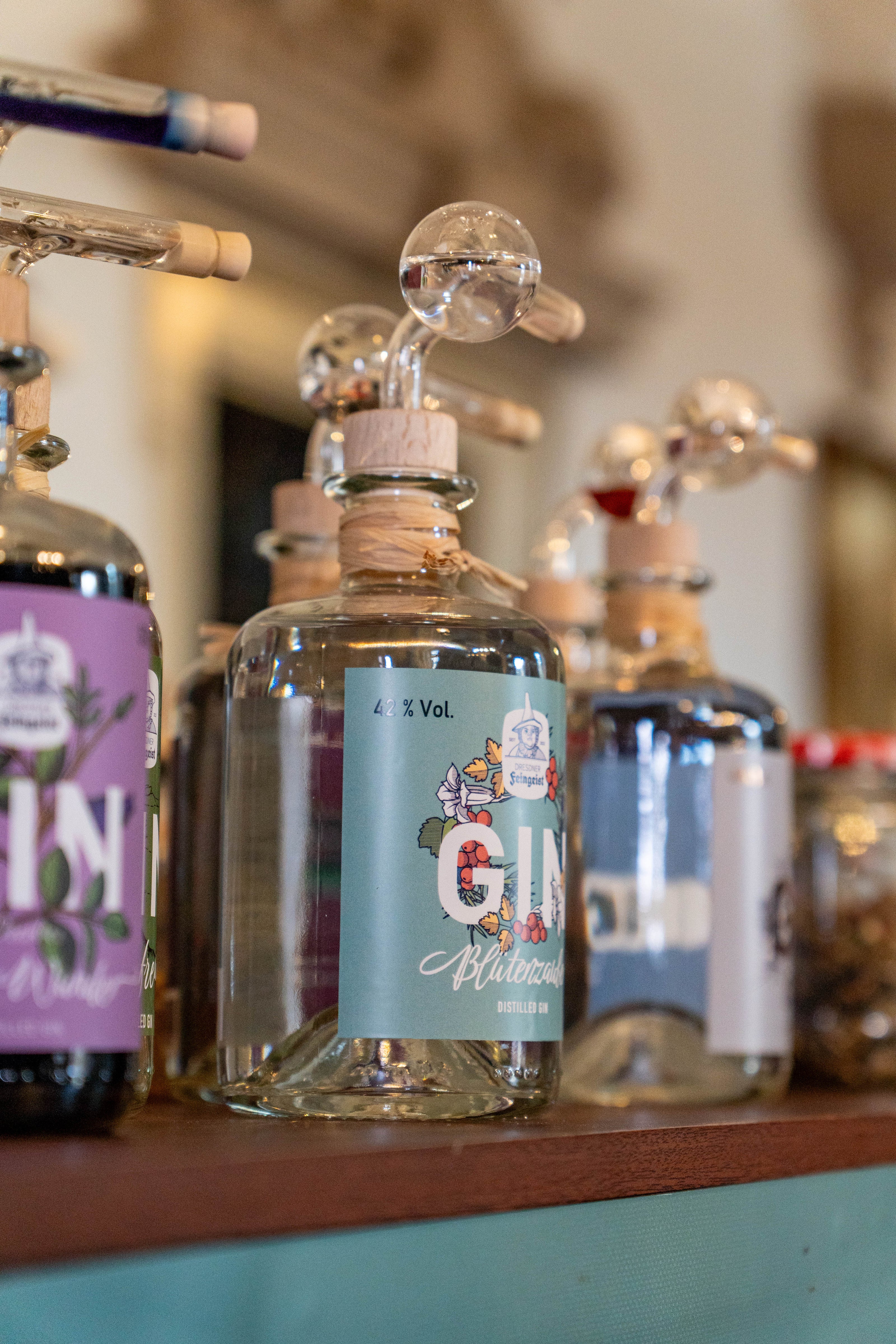 Verschiedene Ginflaschen von Dresdner Feingeist stehen aufgereiht auf einem Tresen. Im Fokus steht die Sorte „Blaue Wunder“ mit floraler Etikettgestaltung und 42 % Vol., ausgestattet mit einem Probieraufsatz aus Glas. Links daneben ist eine lila Flasche „Classic Gin“ zu erkennen, rechts weitere Sorten leicht unscharf im Hintergrund. Die Szene wirkt wie auf einem Verkostungsevent oder einem Marktstand.