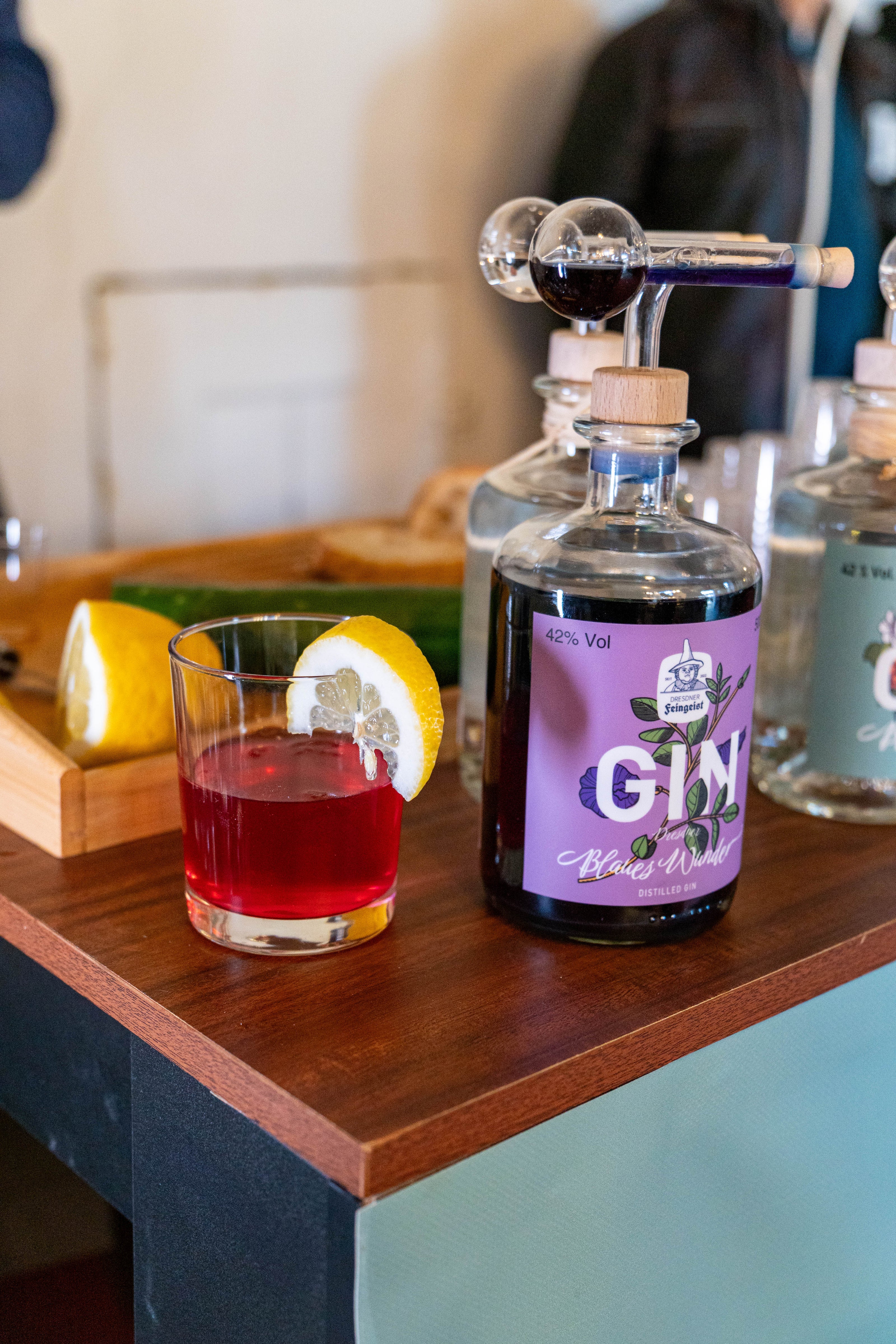 Eine geöffnete Flasche „Blaues Wunder“ Gin von Dresdner Feingeist steht auf einem Tresen. Daneben ein Cocktailglas mit einem leuchtend roten Drink, garniert mit einer Zitronenscheibe. Im Hintergrund sind weitere Ginflaschen sowie frische Zitronen und Gurkenstücke zu sehen – Teil einer Verkostungsszene.