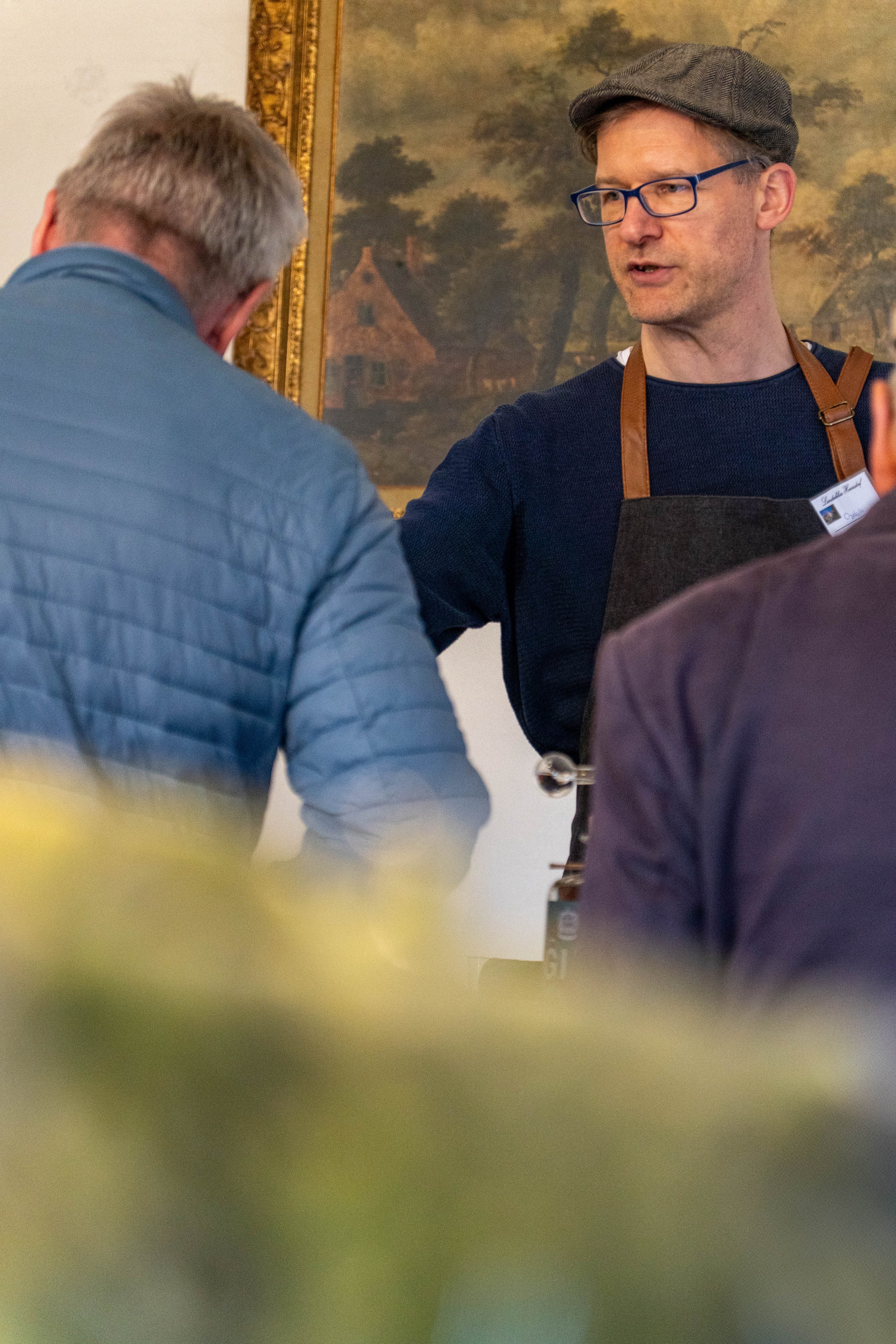 Ein Mann mit Schiebermütze, Brille und dunkler Schürze steht hinter einem Tresen auf einem Event. Er erklärt etwas einem Besucher, der mit dem Rücken zur Kamera steht. Im Hintergrund ist ein großes, gerahmtes Gemälde zu sehen. Es handelt sich um eine Szene vom Dresdner Feingeist Stand beim Hoffest.