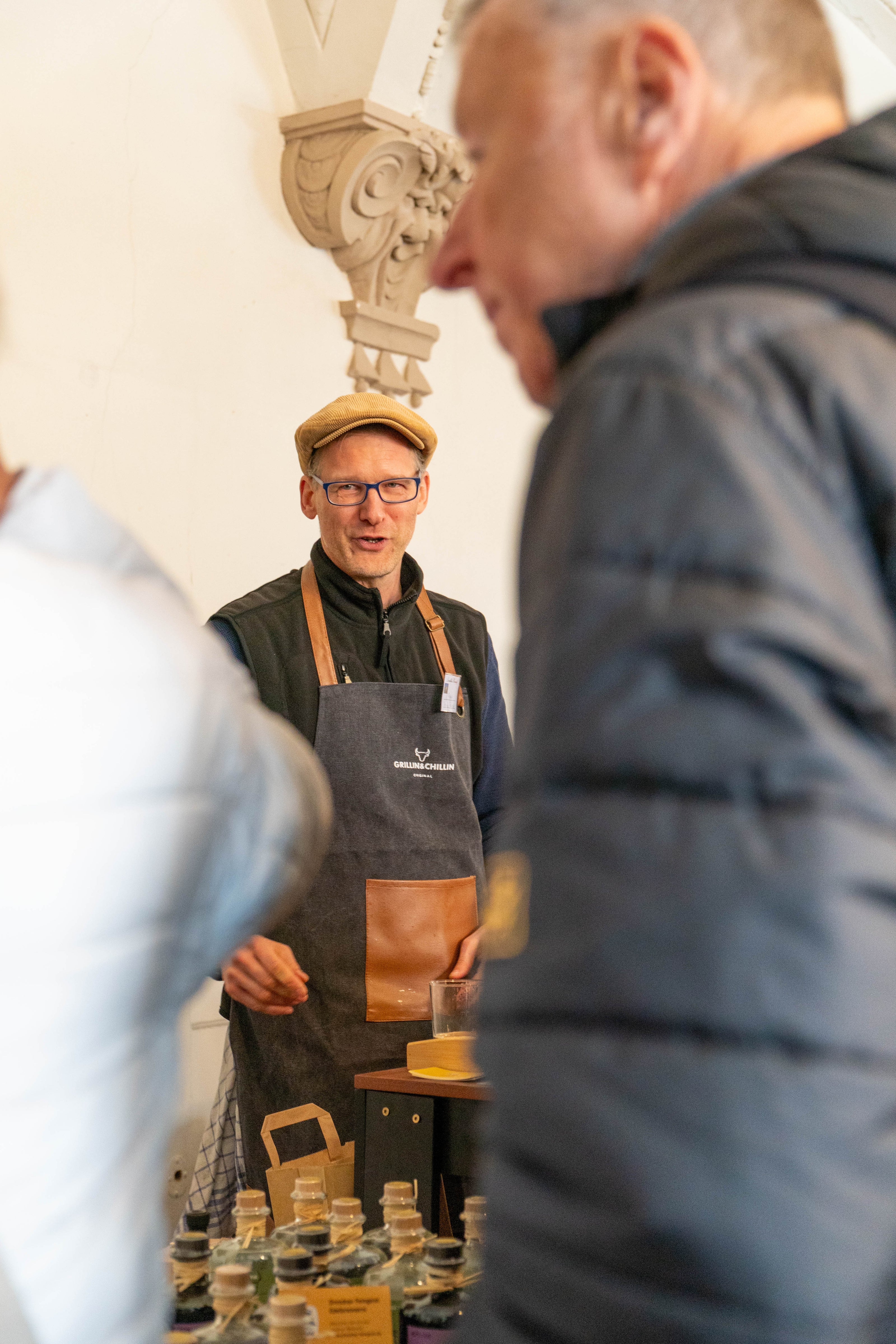 Ein Mann mit Schiebermütze, Brille und dunkler Schürze steht hinter einem Tresen auf einem Event. Er erklärt etwas einem Besucher, der mit dem Rücken zur Kamera steht. Im Hintergrund ist ein großes, gerahmtes Gemälde zu sehen. Es handelt sich um eine Szene vom Dresdner Feingeist Stand beim Hoffest.