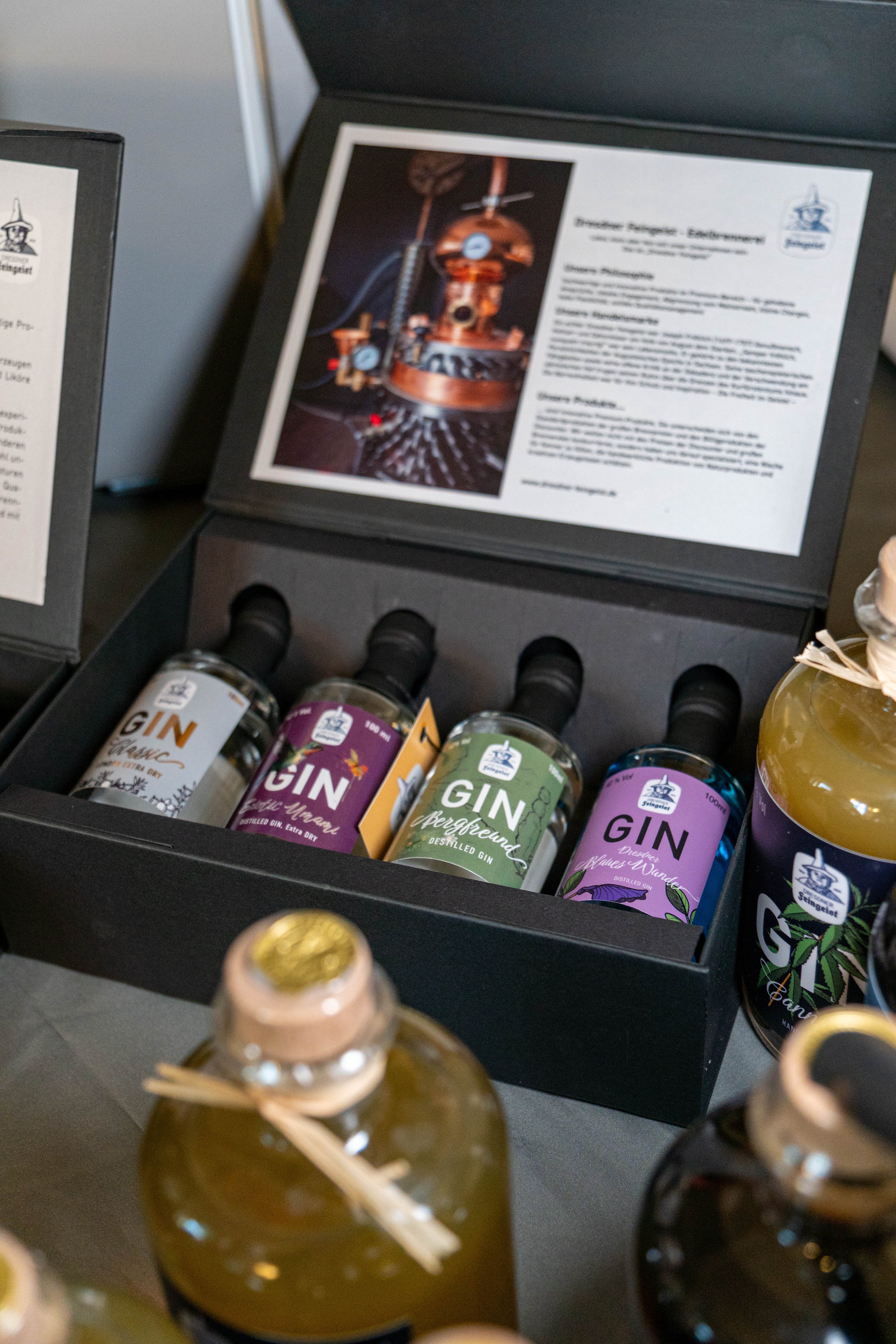 Offene Tastingbox von Dresdner Feingeist mit vier Mini-Ginflaschen: Classic, Exotic Umami, Bergfreund und Blaues Wunder. Im Deckel befindet sich eine Informationstafel mit Bild der Kupferdestille. Daneben stehen größere Flaschen, u. a. der Cannabis Gin.