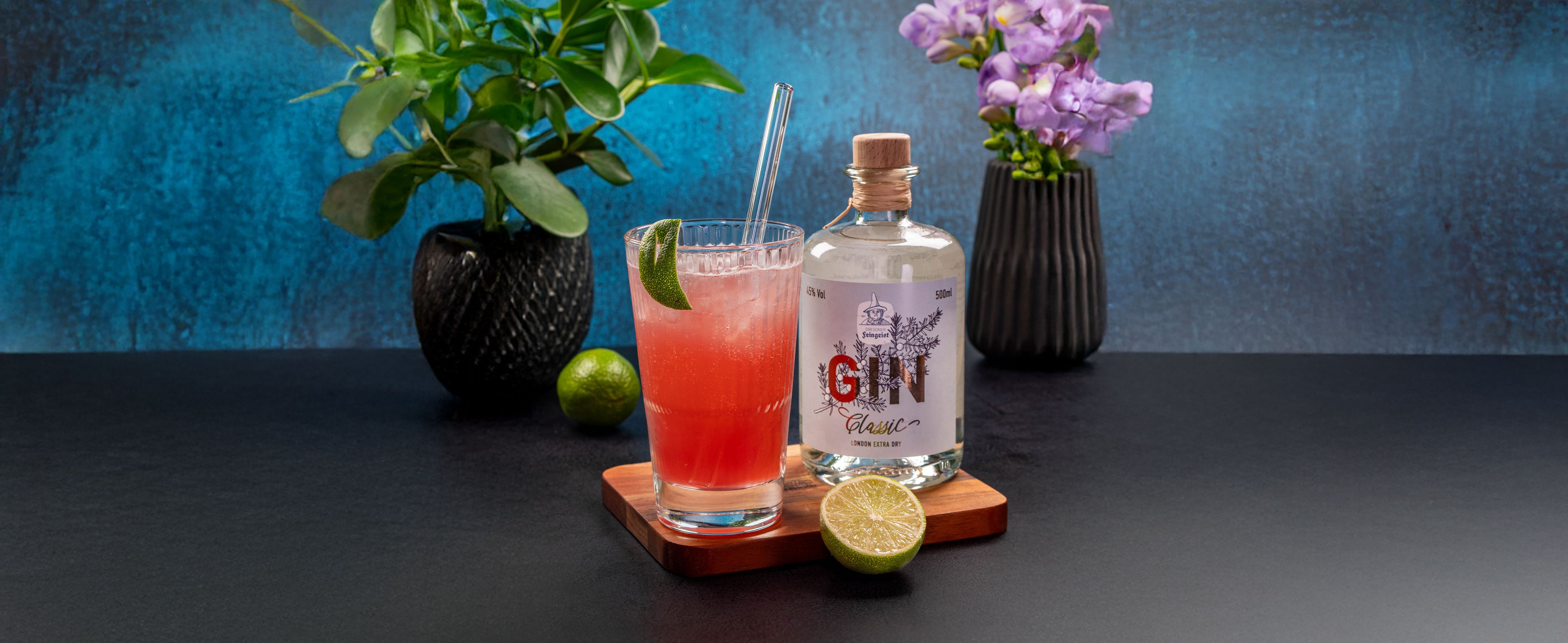 Frischer Cocktail mit Dresdner Feingeist Classic Gin, serviert in einem hohen Glas mit Strohhalm und Limettenscheibe. Daneben eine halbierte Limette und die Ginflasche auf einem Holzbrett. Im Hintergrund zieren Pflanzen und lilafarbene Blumen die Szene auf dunklem Untergrund mit leuchtend blauem Hintergrund.