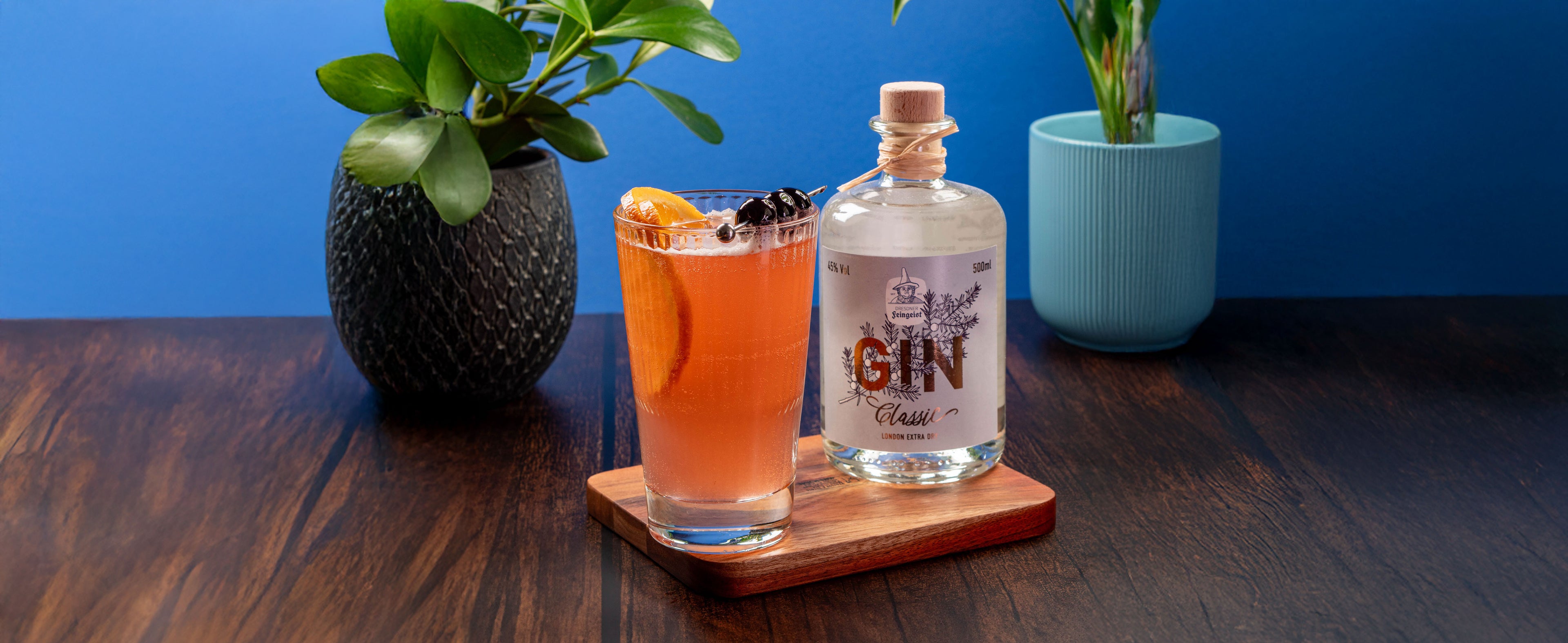Erfrischender Singapore Sling mit Orangenscheibe und Cocktailkirschen in einem hohen Glas, serviert auf einem Holzbrett neben einer Flasche Dresdner Feingeist Classic Gin. Im Hintergrund stehen zwei moderne Blumentöpfe mit grünen Pflanzen auf einem dunklen Holztisch vor blauem Hintergrund.