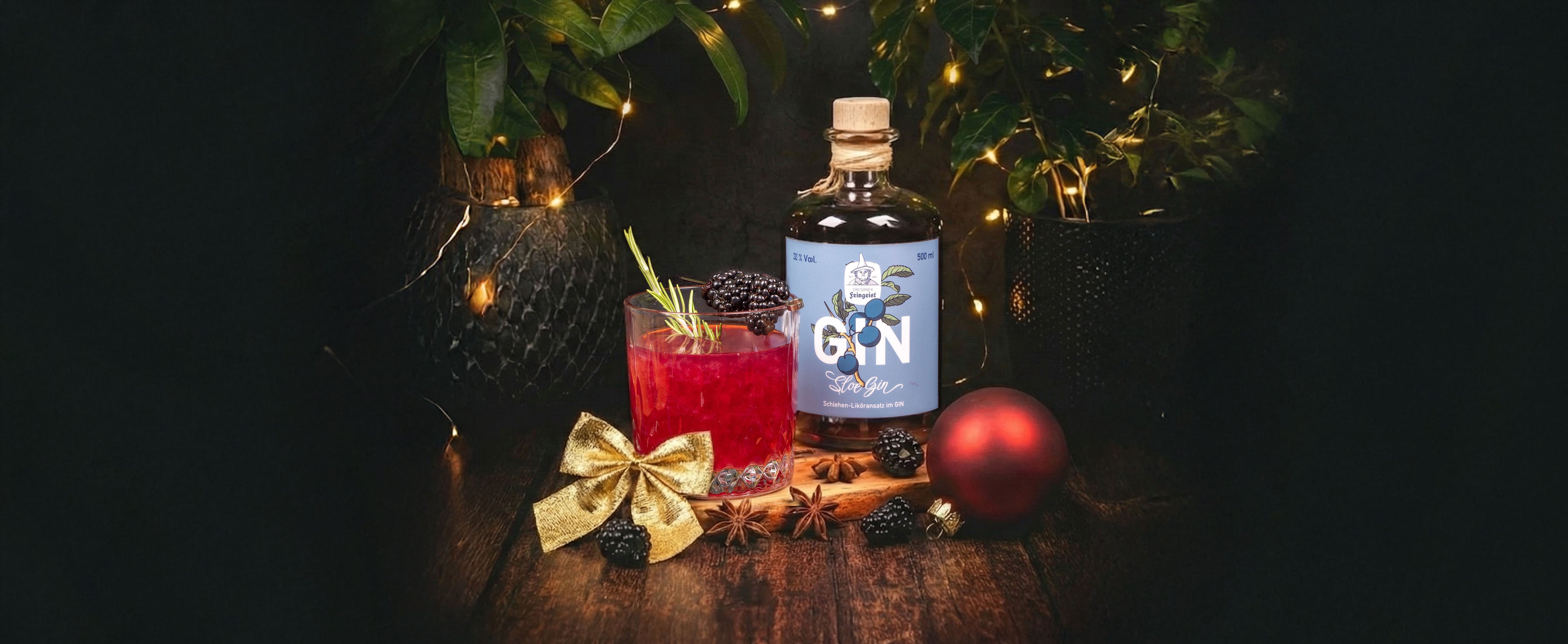 Roter Gin-Cocktail im Tumbler mit Rosmarinzweig und dunklen Beeren vor einer Flasche Dresdner Feingeist GIN Classic Blue, ergänzt durch goldene Schleife, Weihnachtskugel, Gewürze und warme Lichter in stimmungsvoller Winterkulisse.