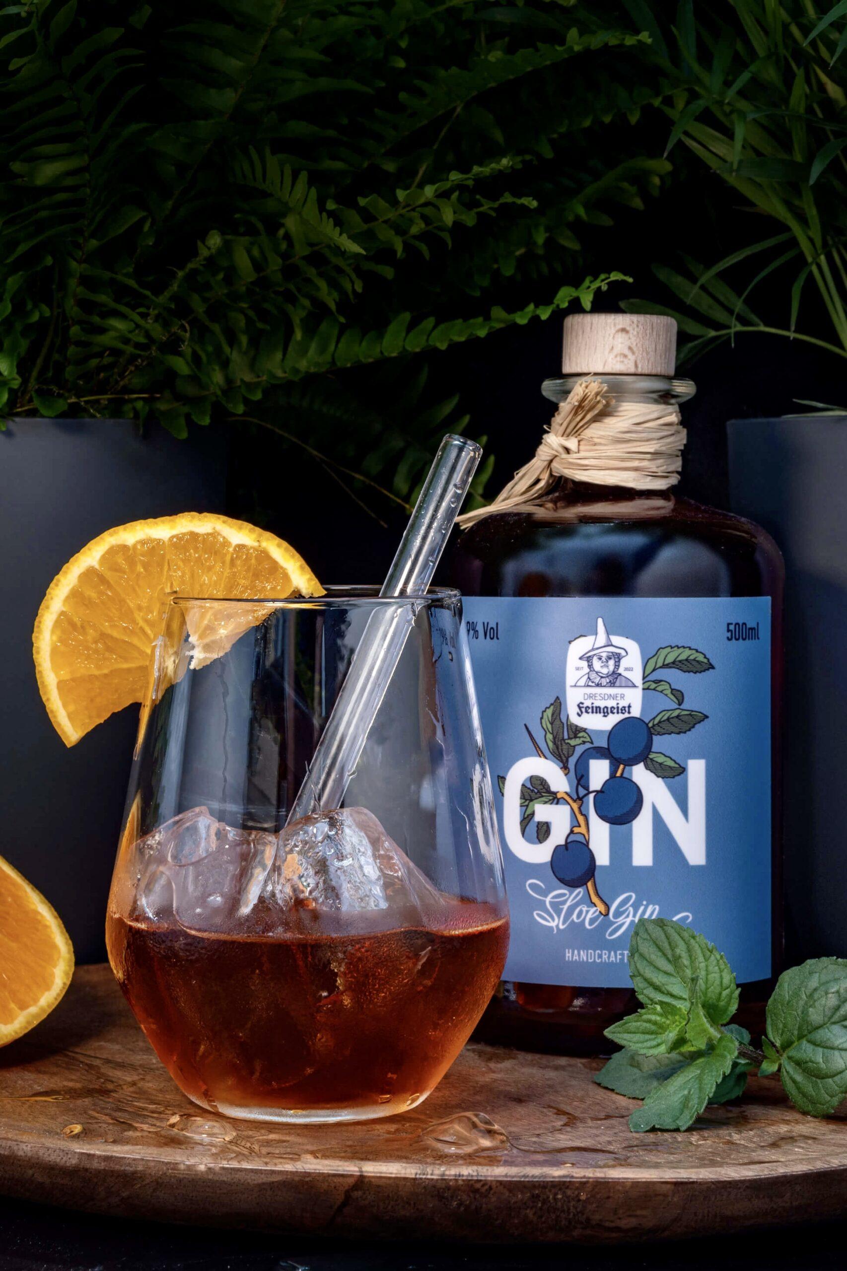Elegante Präsentation des Dresdner Feingeist Sloe Gin: Eine dunkelblaue Flasche steht neben einem Tumbler-Glas mit bernsteinfarbenem Drink, Eiswürfeln, Metallstrohhalm und Orangenscheibe. Umgeben von Minze, Zitrone und dunklem Hintergrund mit Farnen entsteht eine edle, natürliche Atmosphäre.