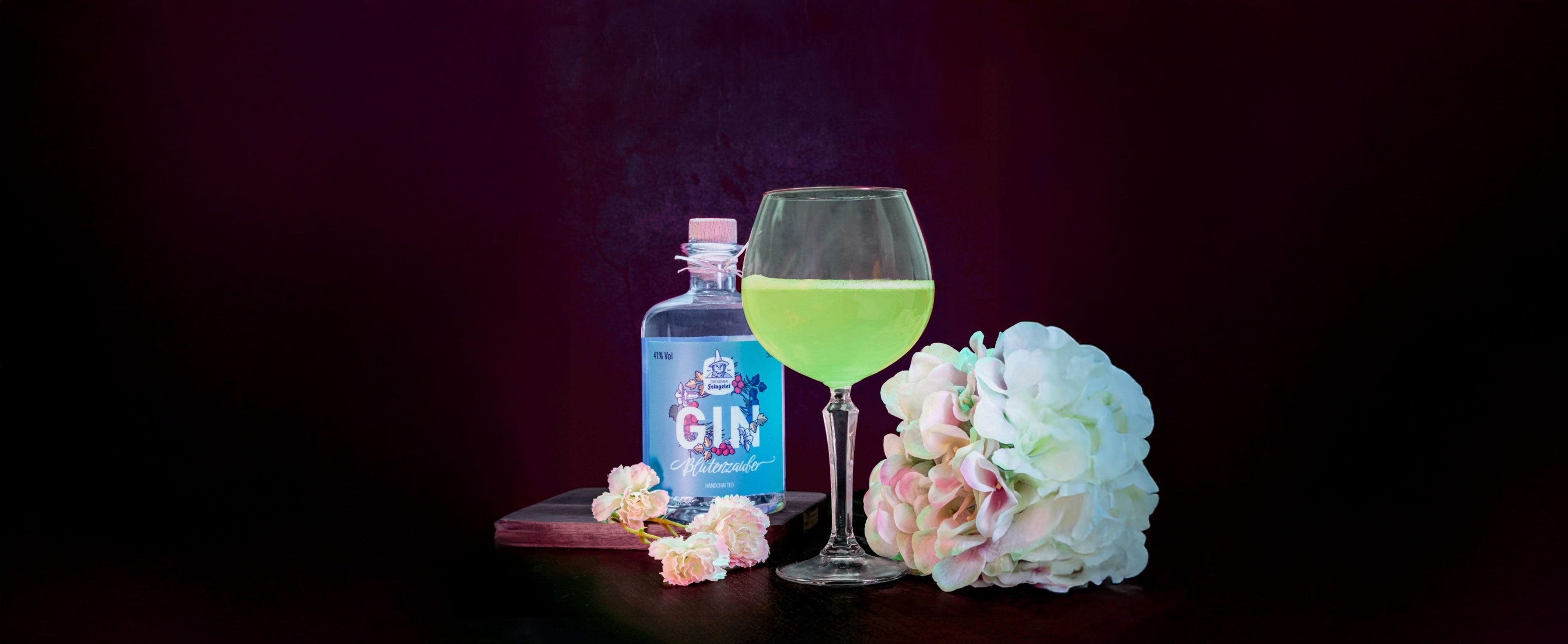 Ein leuchtend grüner Cocktail in einem bauchigen Weinglas steht auf einem dunklen Tisch neben einer Flasche Dresdner Feingeist Blütenzauber Gin. Weiße und zartrosa Blüten umrahmen das Arrangement, das vor einem tief dunkelroten Hintergrund stilvoll in Szene gesetzt ist.