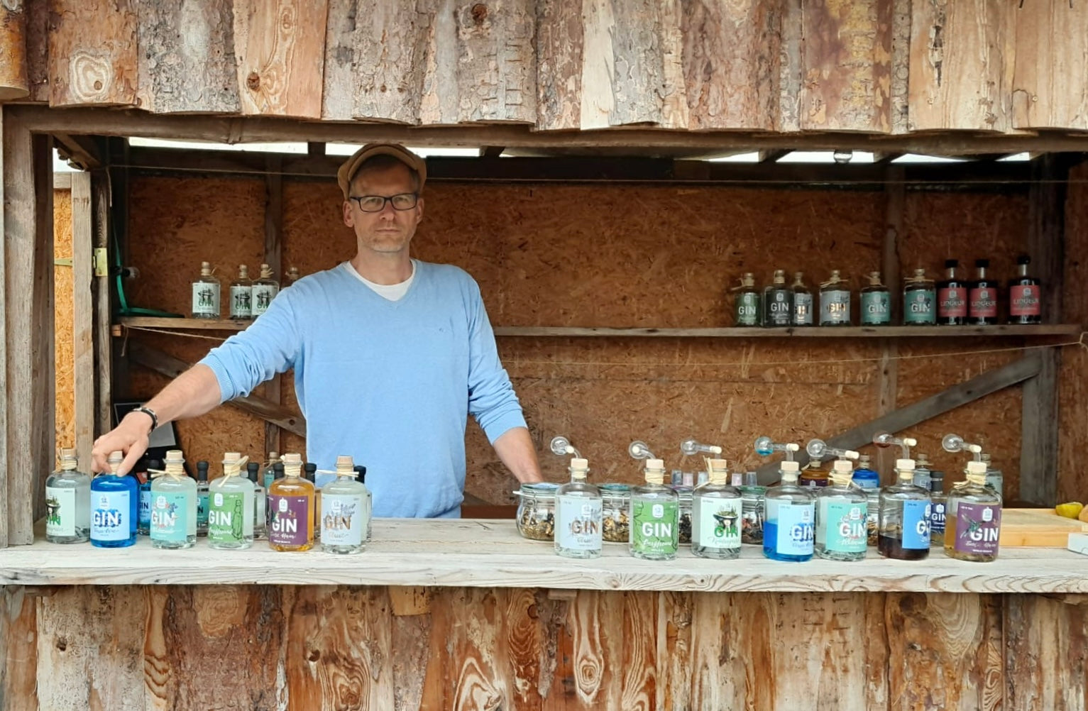 Mann steht hinter einem rustikalen Holzverkaufsstand und präsentiert eine Auswahl verschiedener Dresdner Feingeist Gin-Flaschen, ordentlich aufgereiht vor ihm, mit weiteren Flaschen im Regal im Hintergrund in natürlicher, handwerklicher Markt-Atmosphäre.
