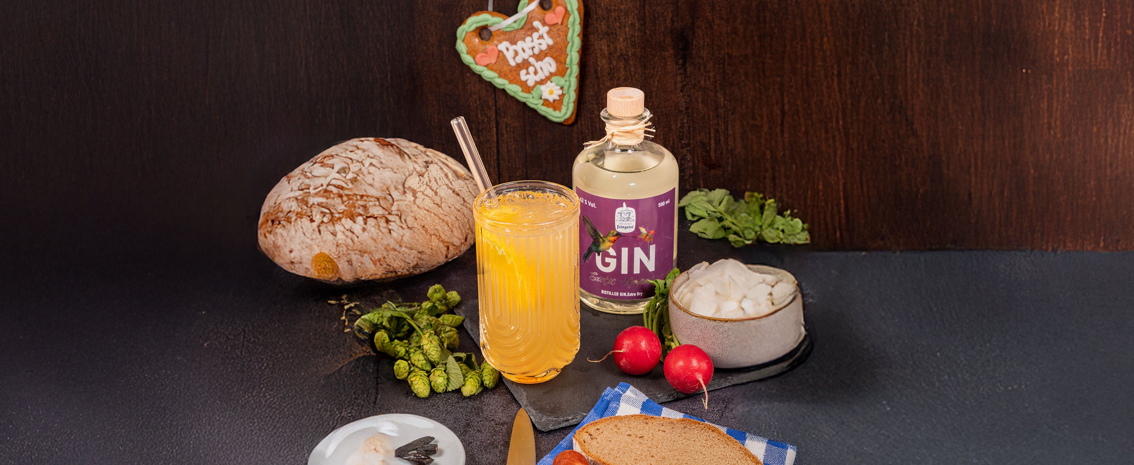 Goldgelber Gin-Longdrink im geriffelten Glas mit Strohhalm neben einer Flasche Dresdner Feingeist GIN Exotic Umami, inszeniert mit Brotlaib, Hopfendolden, Salzschale, Radieschen und rustikalen Elementen in traditionell-herzhafter Atmosphäre.
