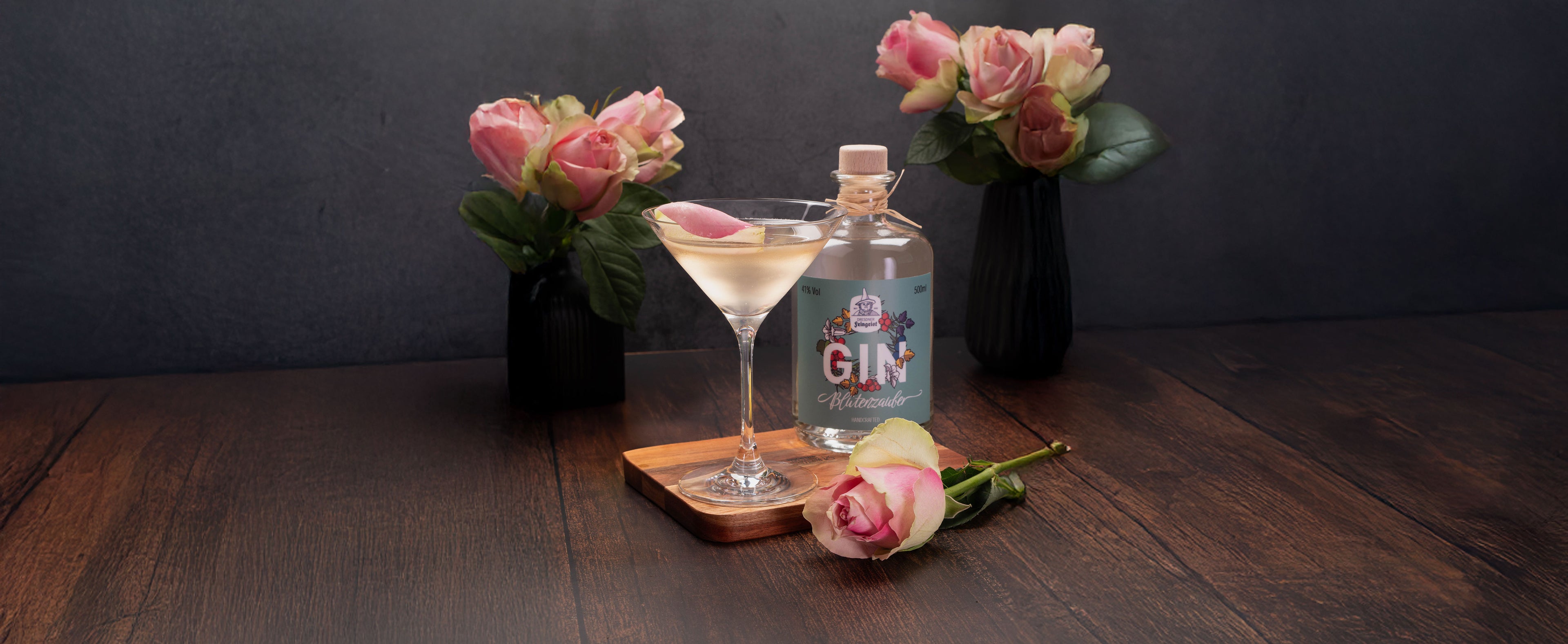Ein eleganter Rose Martini mit Dresdner Feingeist Blütenzauber Gin, garniert mit einem zarten Rosenblatt, arrangiert auf einem Holztisch mit rosafarbenen Rosenvasen im Hintergrund – ein romantisches Setup für den floralen Cocktail.

