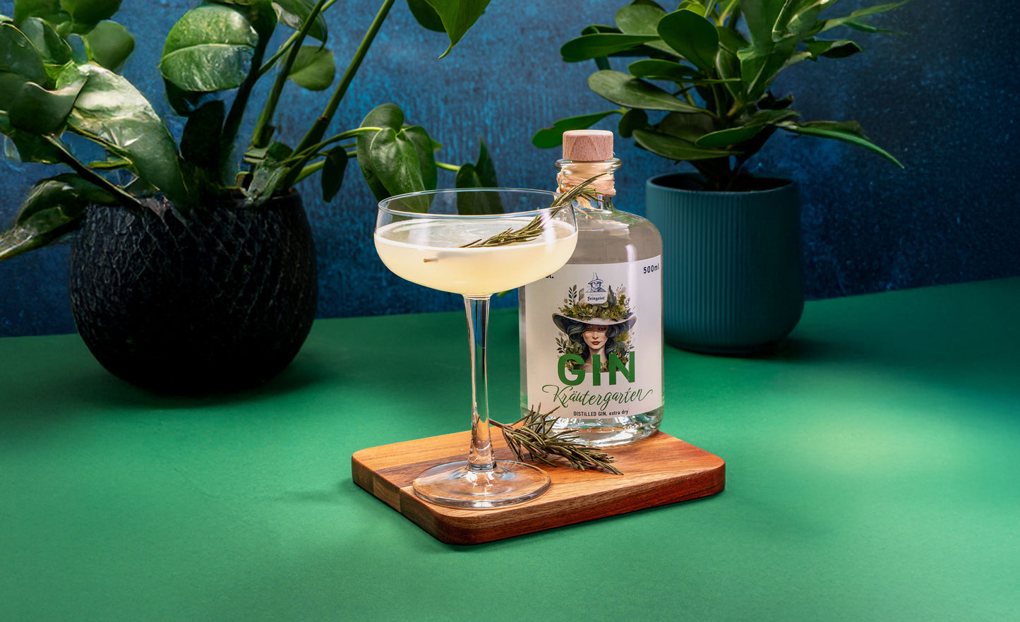 Stimmungsvoll inszenierter Rosmarin Gimlet mit Dresdner Feingeist Gin Kräutergarten, serviert auf Holzbrett mit Rosmarinzweig, vor tropischer Pflanzenkulisse.