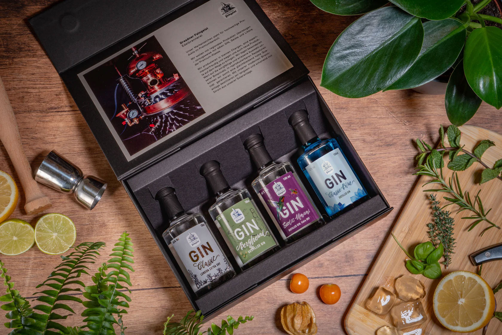 Tastingbox mit vier 100 ml-Flaschen Dresdner Feingeist Gin (Classic, Bergfreund, Exotic Umami und Classic Blue) in schwarzer Geschenkverpackung, umgeben von frischen Zutaten und Bar-Tools auf einem Holztisch.